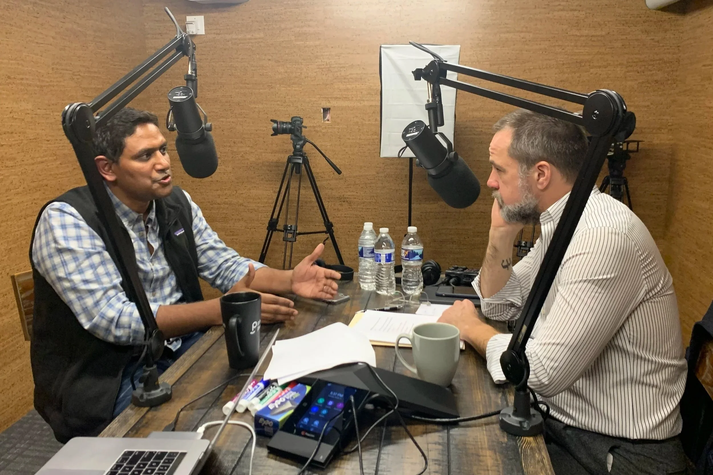 Two men are having a conversation in a recording studio with microphones, cameras, water bottles, and recording equipment on the table. Derek Herman, communications executive, set up this media interview with a CEO and the podcast reporter.