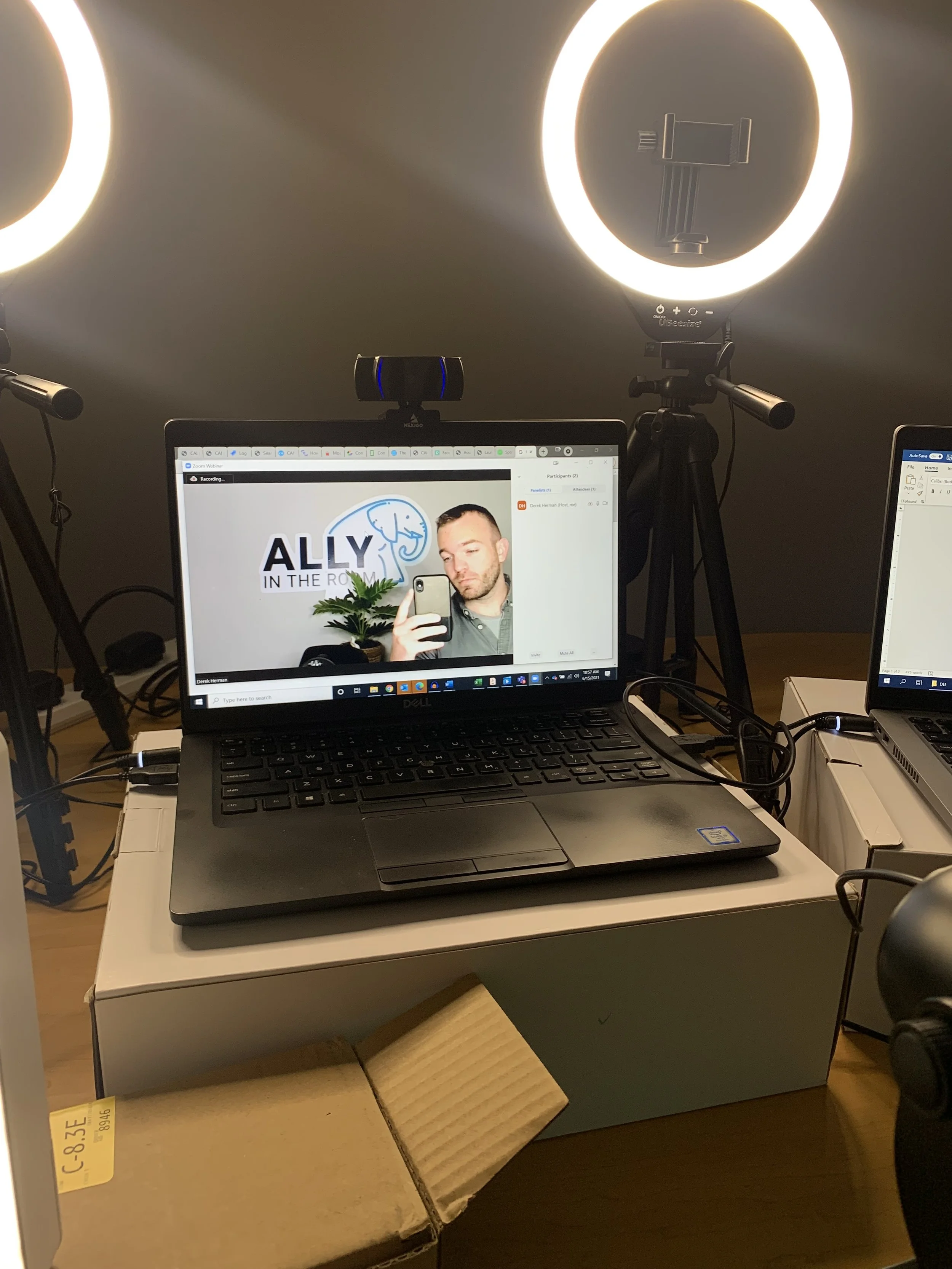A workspace setup with a laptop on a white box, two ring lights illuminating the area, a webcam, and a smartphone attached to a tripod. The laptop screen displays a man taking a selfie with a logo that reads 'Ally in the Room' behind him.