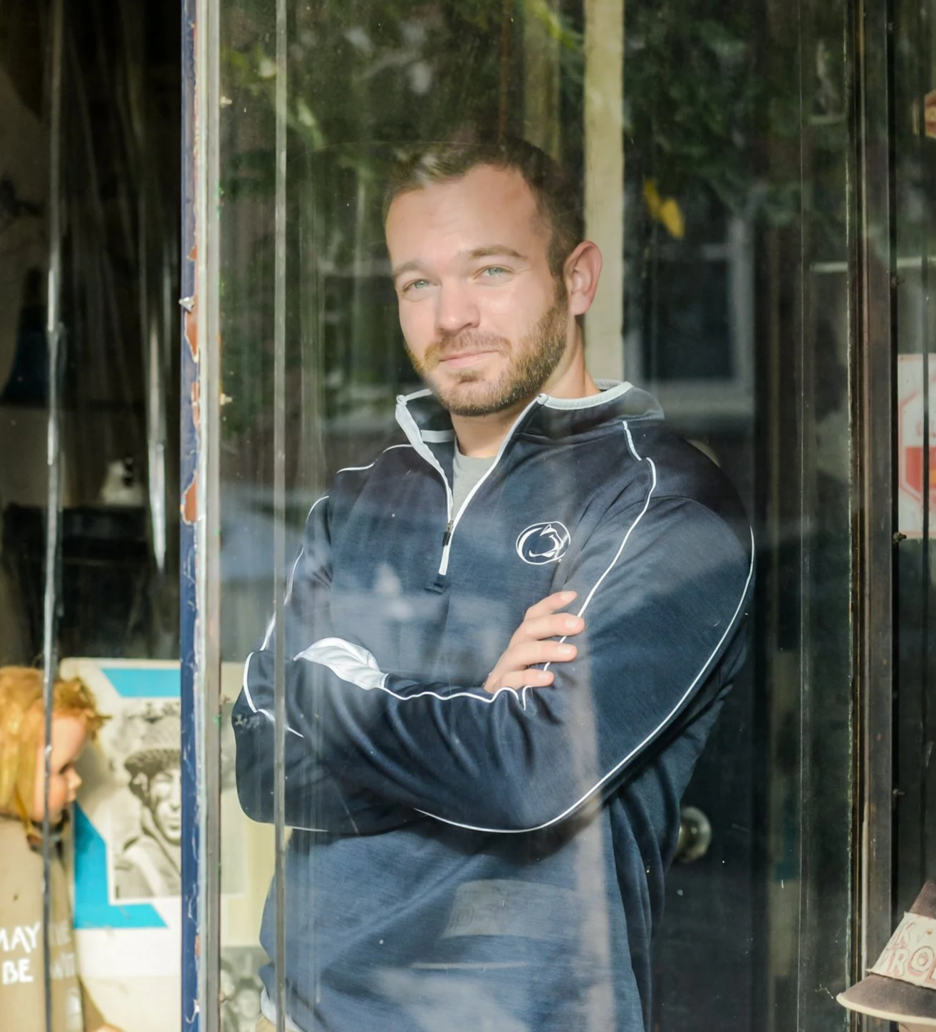 A man with light skin, short brown hair, and a short-cropped beard standing inside a glass display case, arms crossed, wearing a dark athletic jacket with a Penn State logo on the chest. Derek Herman, communications executive.