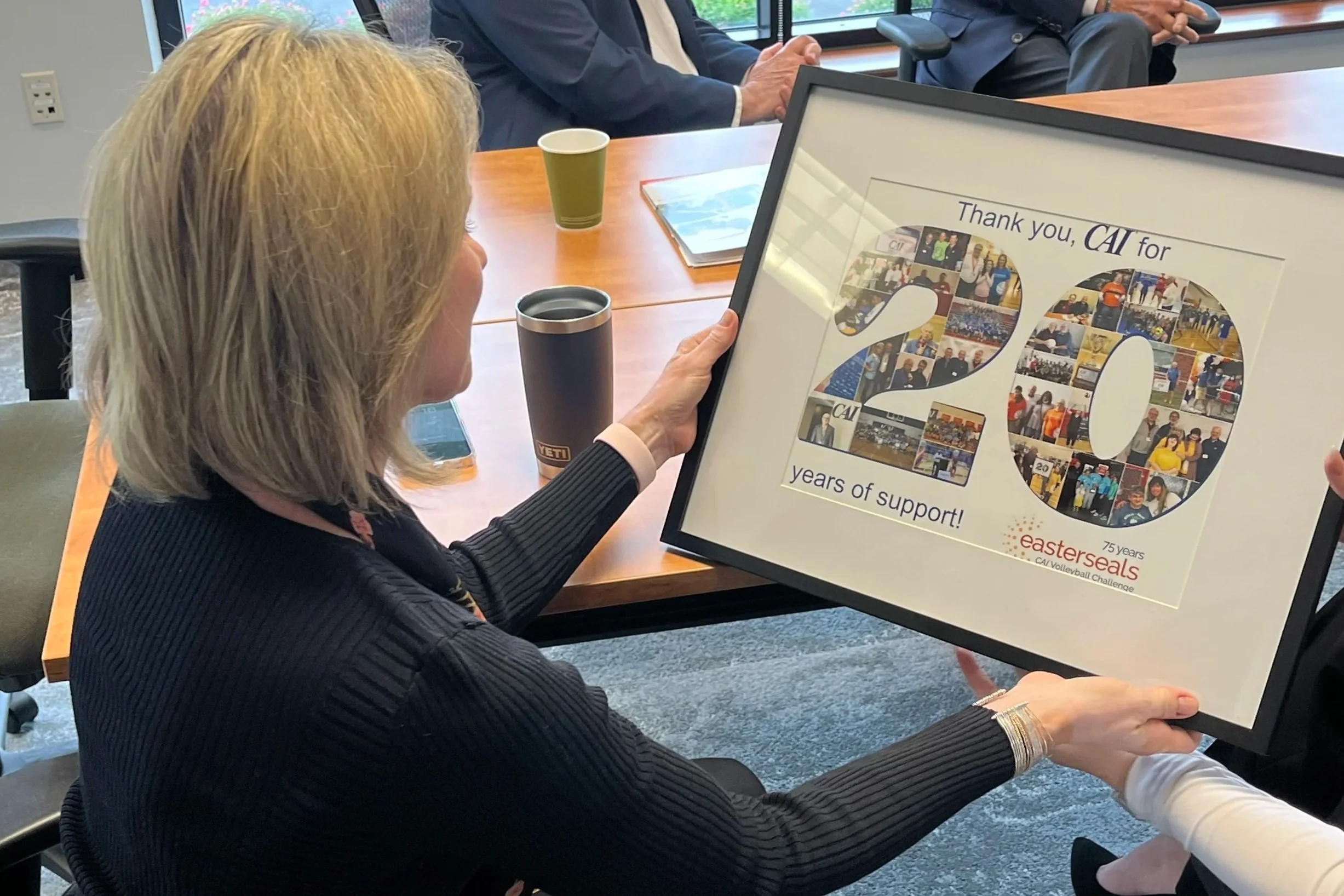 Woman in black jacket holding a framed collage with the number 20 filled with photos, celebrating 20 years of support for CAI, during a meeting with others at the table.