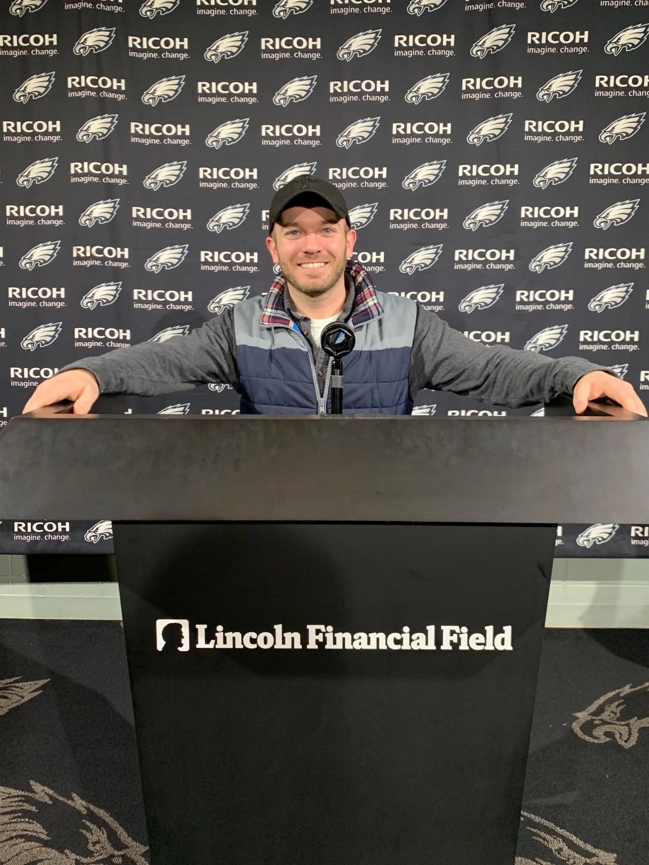 A man smiling seated at a press conference podium with a backdrop featuring the Philadelphia Eagles logo and Ricoh branding at Lincoln Financial Field. This is Derek Herman, a communications executive, specializing internal and external communication