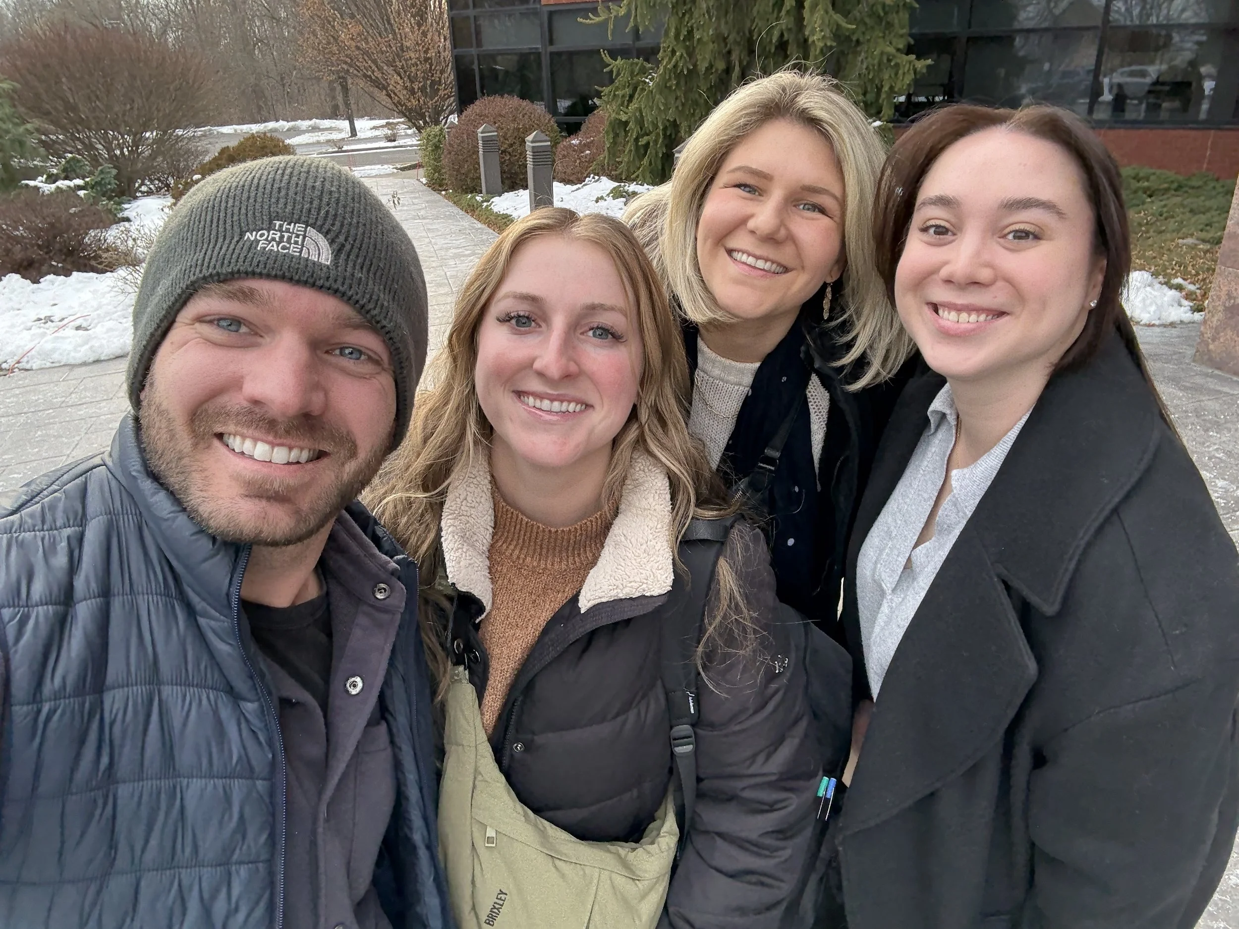 Four smiling people taking a selfie outdoors in winter with snow and trees in the background. This is Derek Herman, communications executive, and his team whose skills span internal communications, PR, and employer branding.