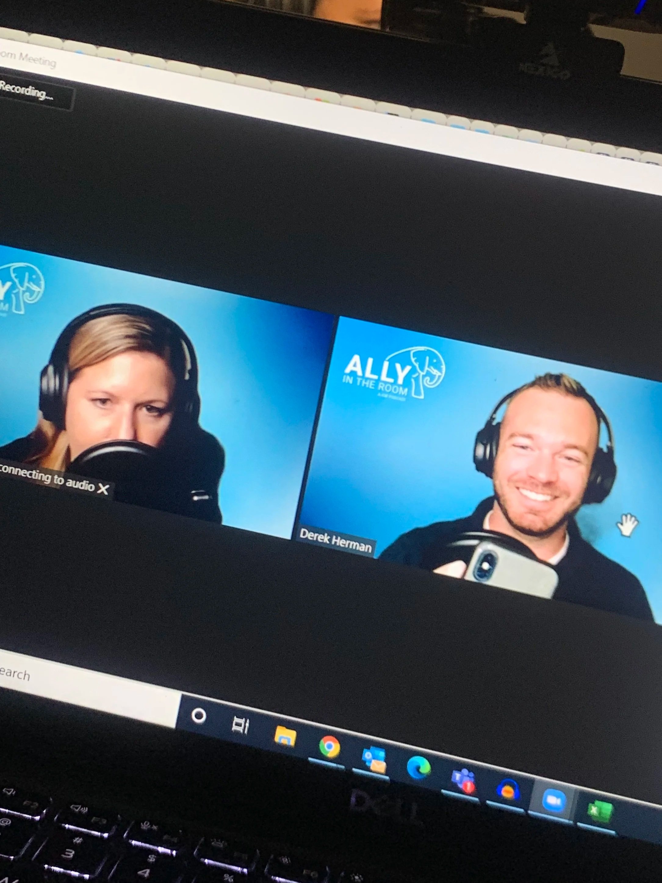 Screenshot of a virtual meeting with two participants, a woman with headphones speaking into a microphone and a smiling man with headphones holding a phone, with logos and text indicating 'Ally in the Room' and the man's name, Derek Herman.