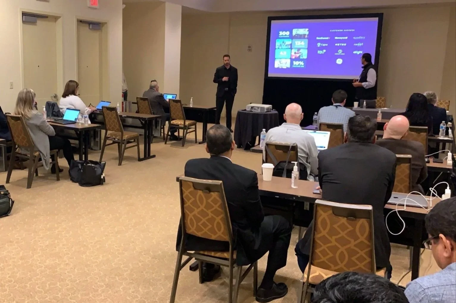 A group of people attending a business presentation or conference in a conference room. The presenter is standing near a large screen displaying a slide, with attendees seated at tables using laptops and tablets.