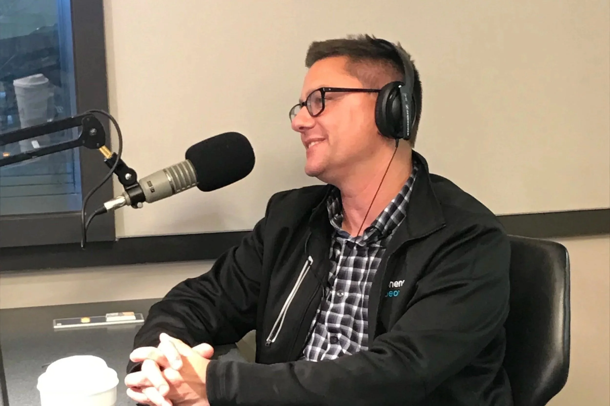A man wearing glasses and a black jacket, sitting at a table with clasped hands, speaking into a microphone in a recording studio.