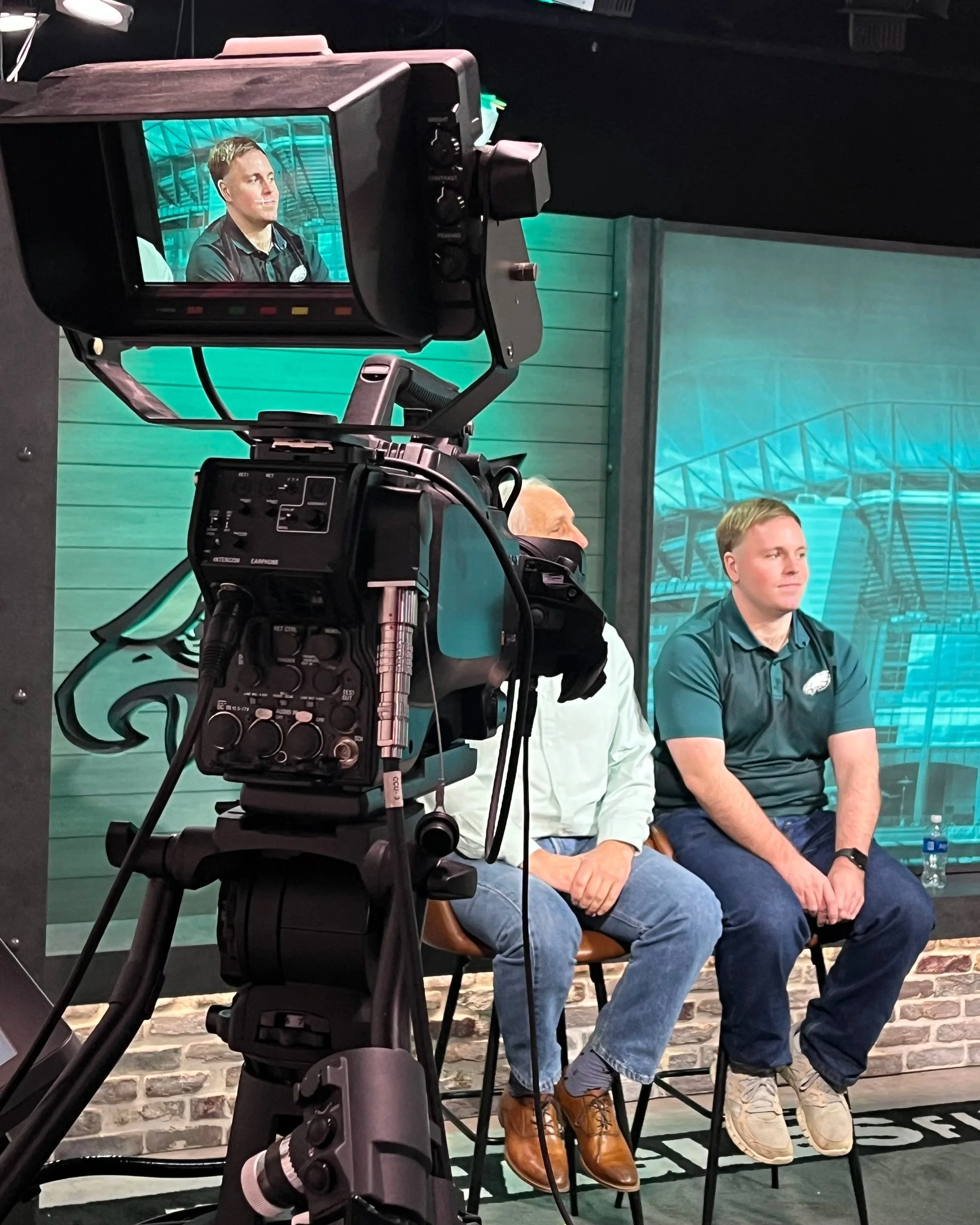 Two men sitting on chairs in front of a teal and gray mural of the Philadelphia Eagles football team, with a camera recording them during a video or interview.