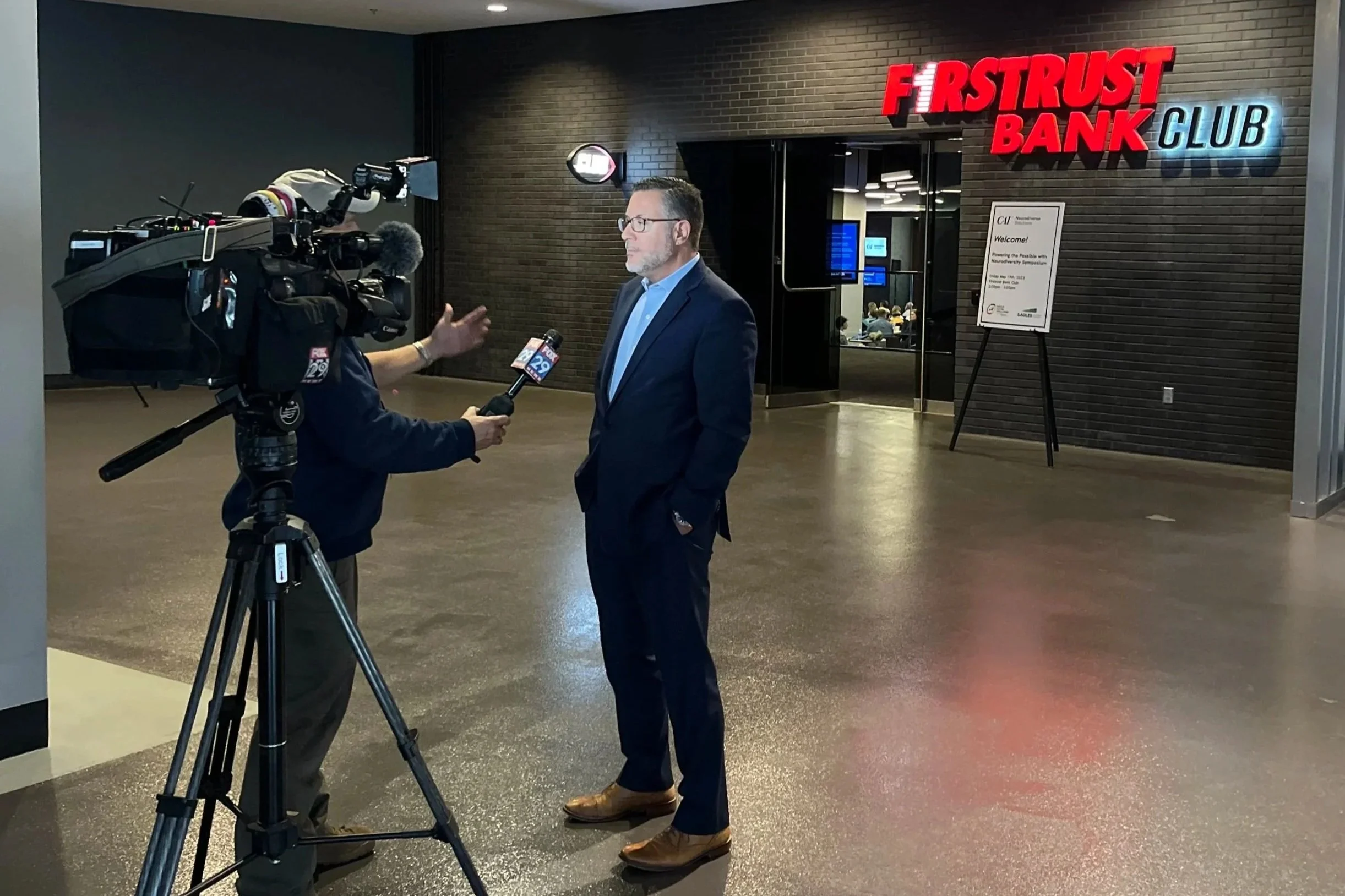 A man in a dark blue suit and blue shirt giving a media interview to a news reporter holding a microphone inside a lobby area of First Trust Bank Club. There is a signboard and a hallway behind them.