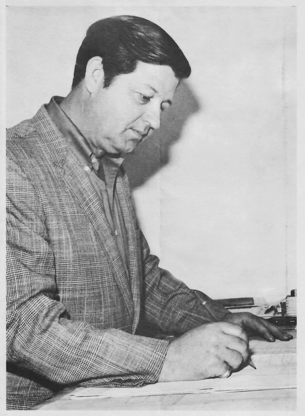 A black-and-white photo of Jim Henry, wearing a checkered blazer, writing in a notebook with a pen, seated at a desk.