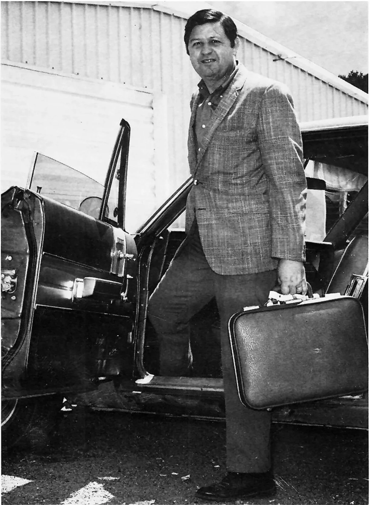 Jim Henry in a checkered blazer and dress pants holding a briefcase standing next to an open vintage car with a suitcase inside. The background shows a building with a sloped roof.