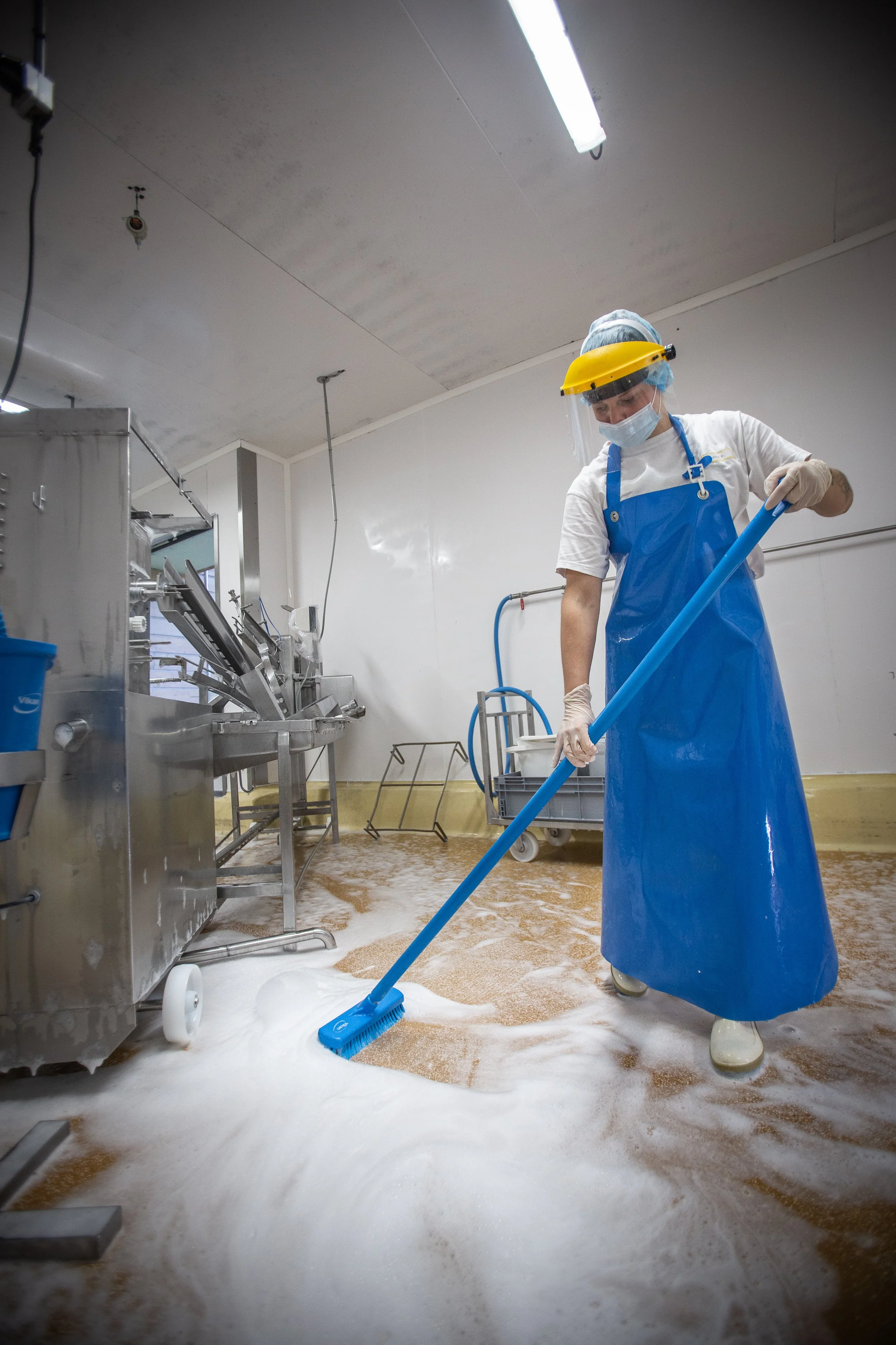 A person wearing protective gear, including a yellow face shield, surgical mask, gloves, and a blue apron, is cleaning a floor with a mop in an industrial or commercial kitchen with foamy soap on the floor.