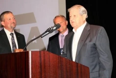 Three men in suits standing behind a wooden podium, with one man speaking into a microphone in front of a plain background.