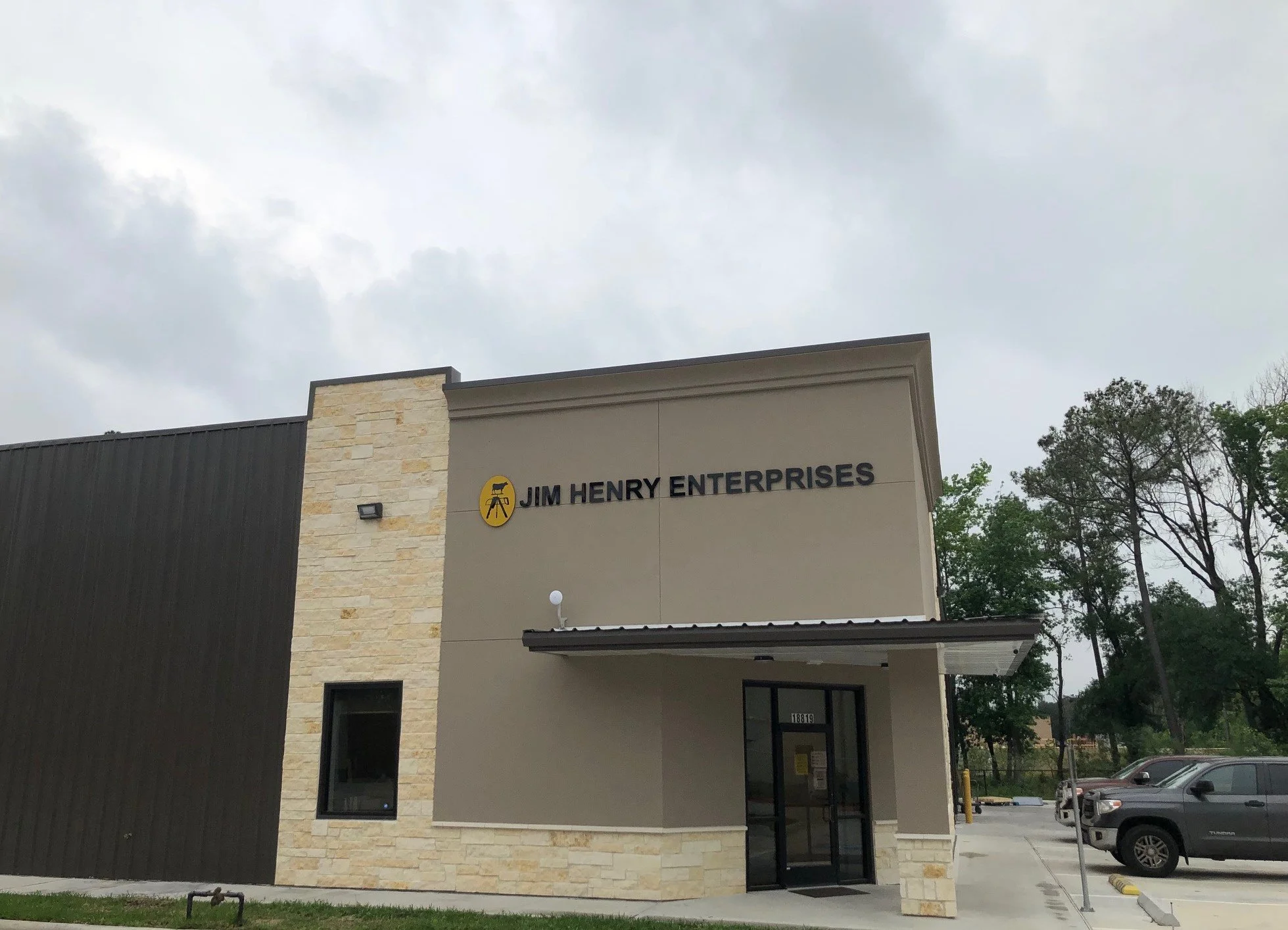 A modern commercial building with stone and dark siding, displaying the sign 'Jim Henry Enterprises' and a logo of a person on a tripod with a camera, under a cloudy sky.