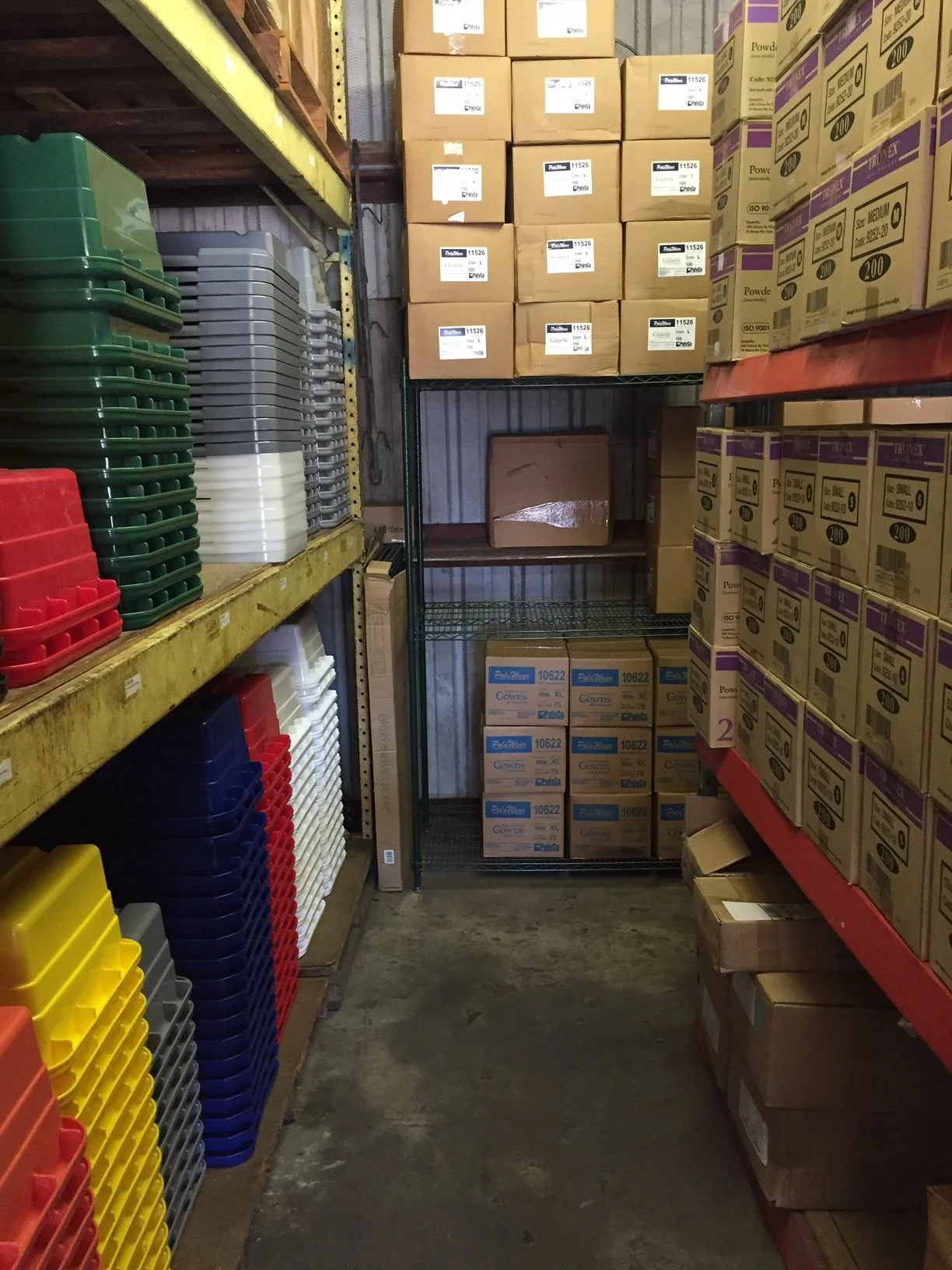 Storage room with organized shelves holding plastic trays in various colors including green, gray, yellow, red, and navy. Cardboard boxes are stacked on shelves and the floor, some labeled with product information, in a warehouse setting.