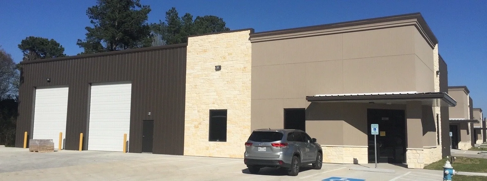 Exterior of a modern industrial building with dark brown and beige walls, a small gray car parked in front, and an accessible parking sign.