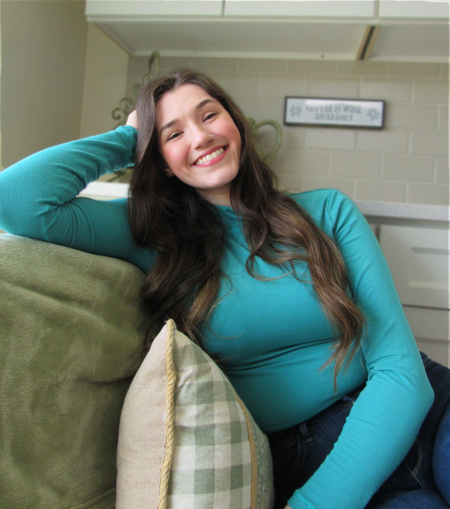 A young woman with long brown hair and a nose ring, smiling, relaxes on a green couch with a beige and green checkered pillow, wearing a teal long-sleeve top.