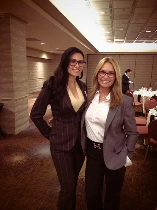Two women in business attire standing in a banquet hall, smiling for the camera.