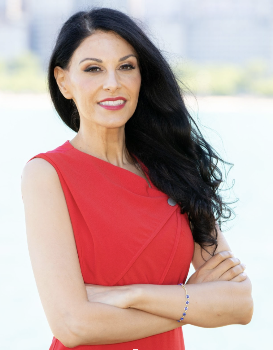 A confident woman with long black hair, wearing a red sleeveless dress and a blue bracelet, standing outdoors with arms crossed.