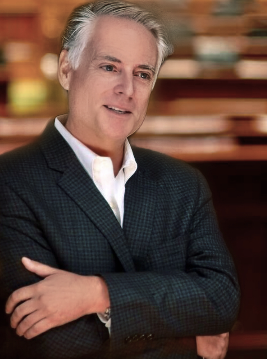 A middle-aged man with gray hair wearing a dark blazer over a white shirt, standing with crossed arms in front of a blurred background.