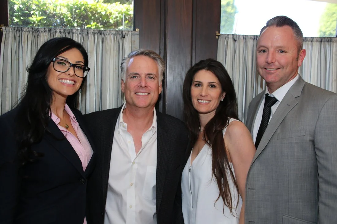 Four people standing indoors near window curtains, smiling at the camera.