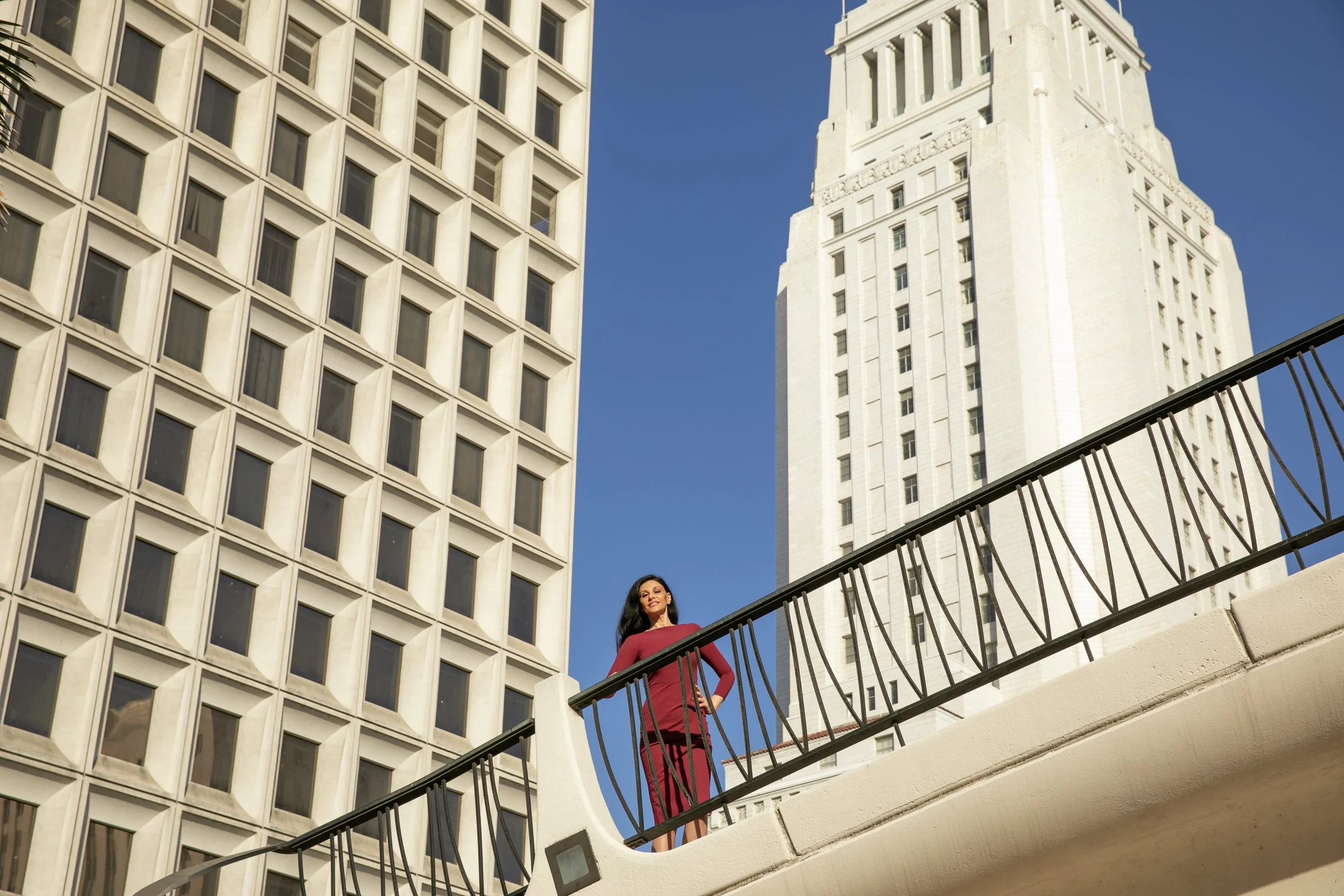 A woman in a red dress standing on an outdoor balcony with a city skyline, tall skyscrapers, and a clear blue sky in the background.