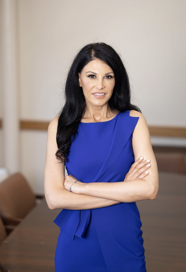 A woman with long black hair wearing a blue sleeveless dress, standing with arms crossed in an indoor setting.