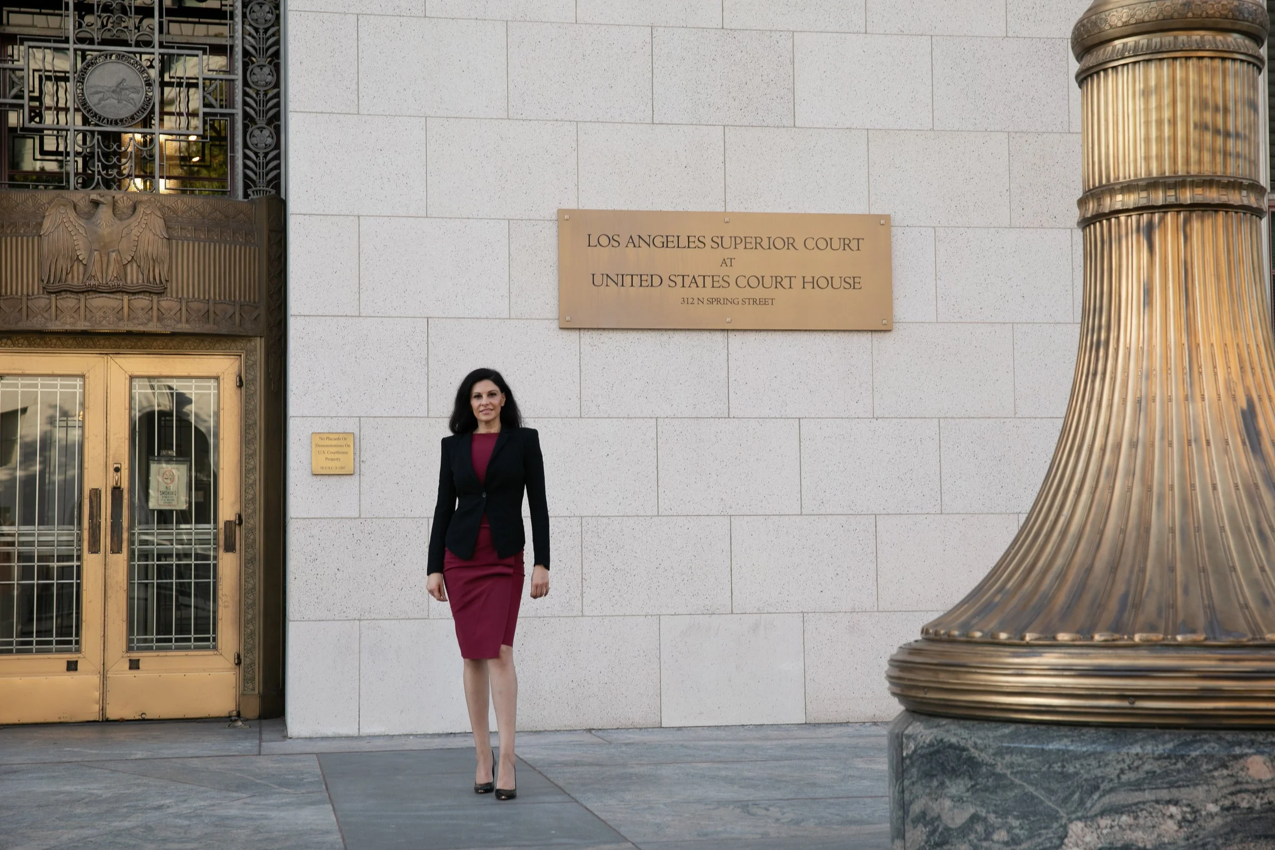 A woman in business attire walking outside the Los Angeles Superior Court at the United States Courthouse, with a large decorative column nearby and a sign on the wall.