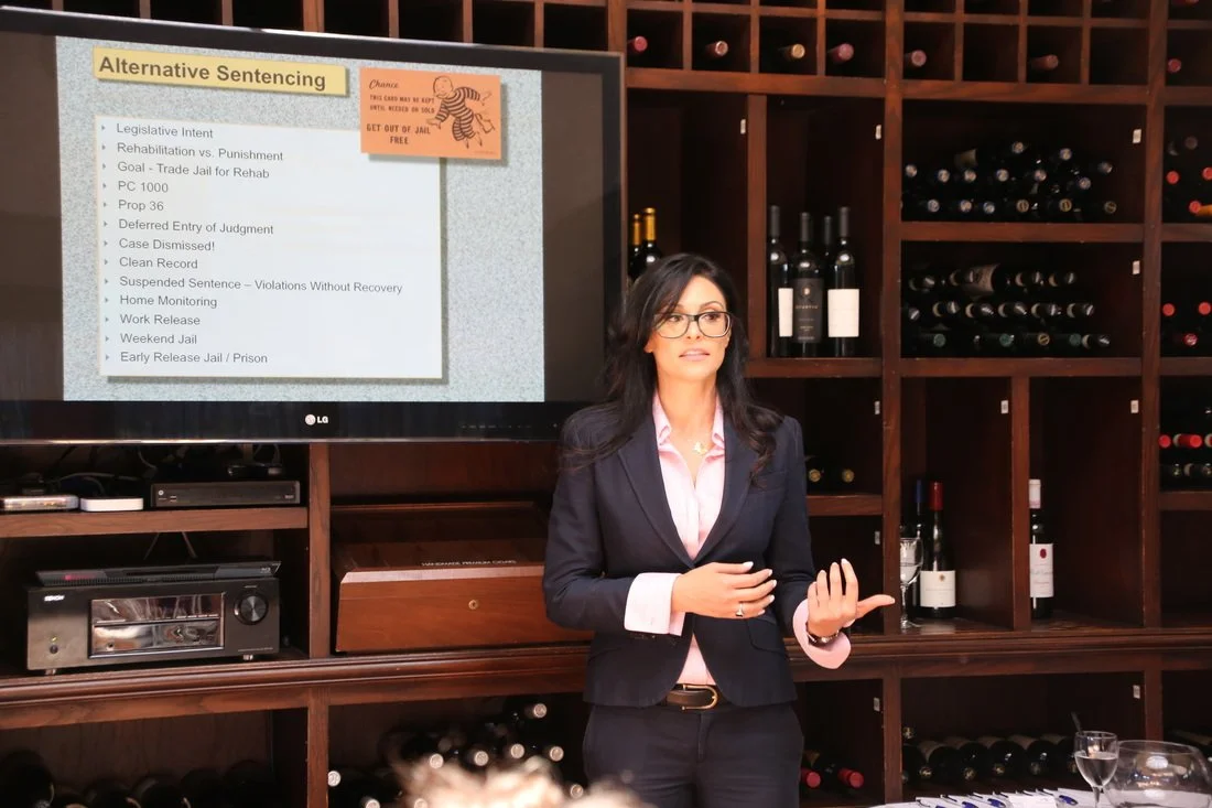 A woman in a dark blazer and glasses giving a presentation in a room with wine bottles on shelves, while a slide on the large screen behind her displays various legal sentences.
