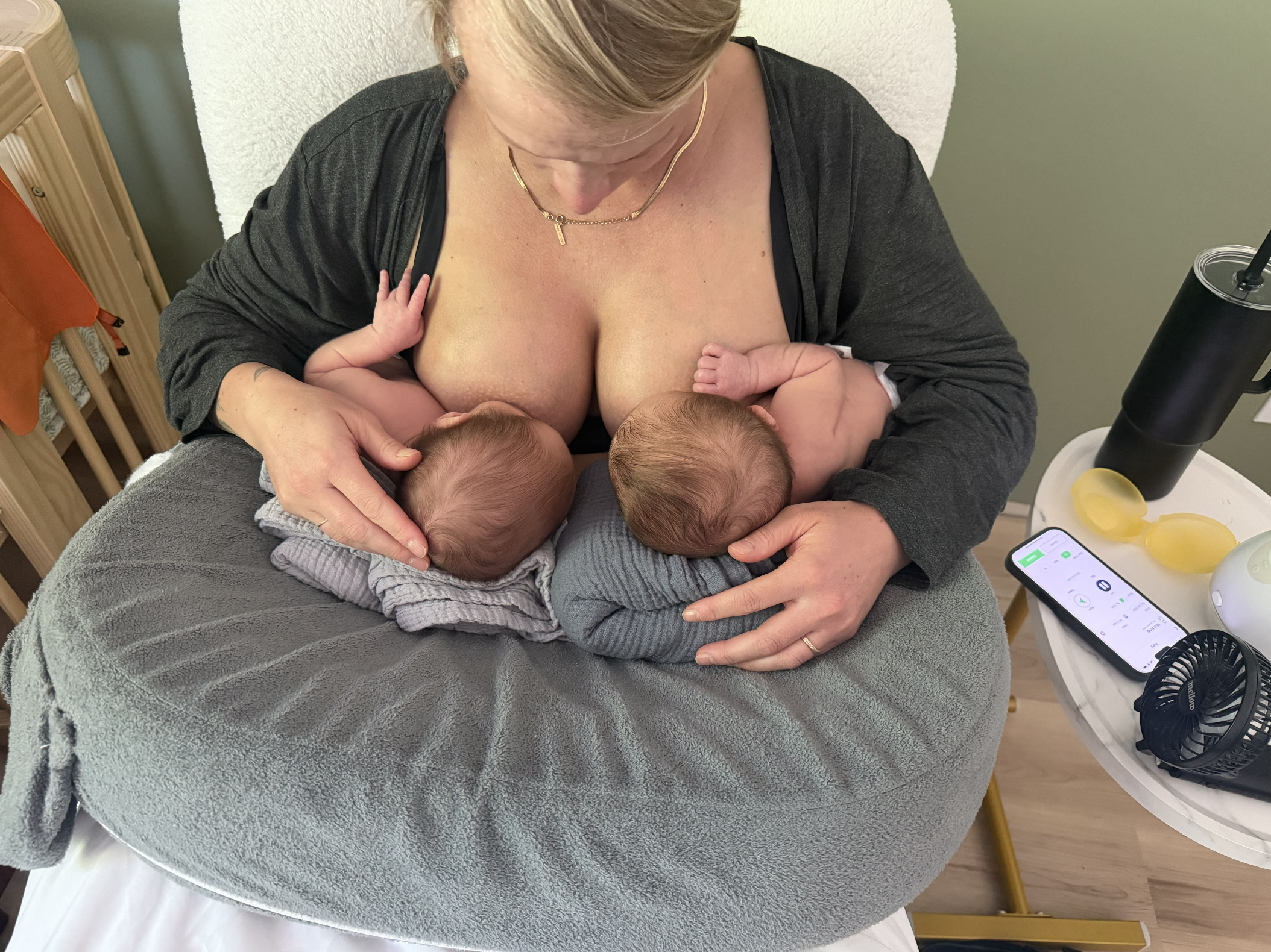 A woman breastfeeding twin babies while sitting on a bed, with a smartphone and some yellow objects nearby.