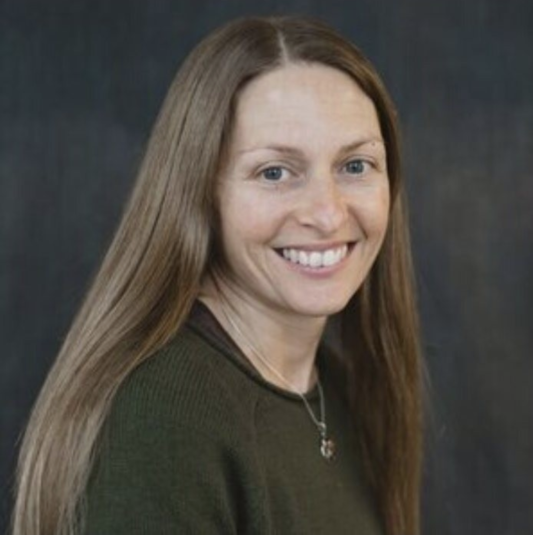 A smiling woman with long reddish-brown hair, wearing a dark green top and a silver necklace, against a dark background.