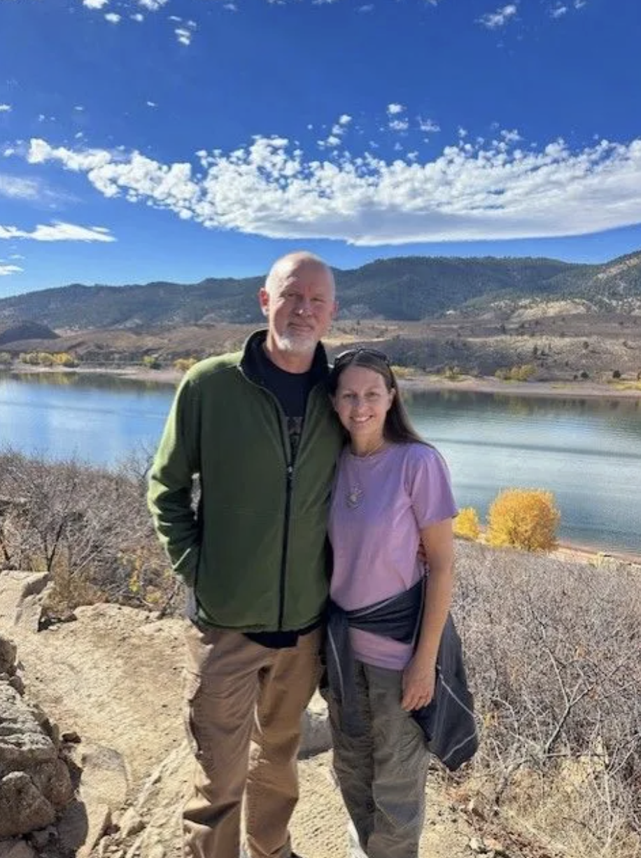 A man and a woman standing side by side outdoors near a body of water with mountains in the background. The sky is blue with white clouds, and there are yellow trees near the shoreline.