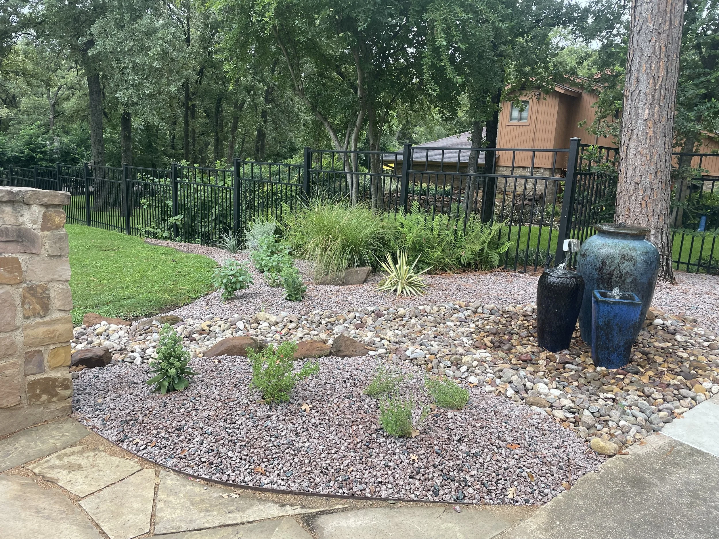A garden with rocks, plants, and two large blue vases, surrounded by a black fence and green trees.