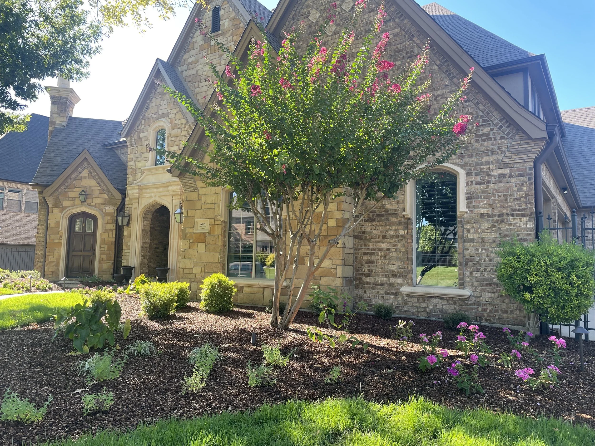 A house with a stone and brick exterior, surrounded by well-maintained landscaping including a tree with pink flowers and various green plants and grass.