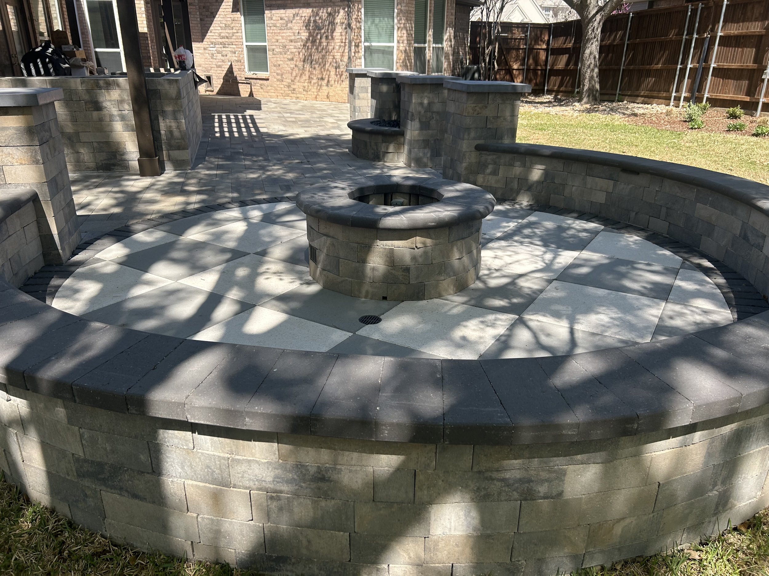 A circular outdoor fire pit with a stone surround, surrounded by a paved patio with shadows of tree branches, in a backyard with a brick house and a wooden fence.