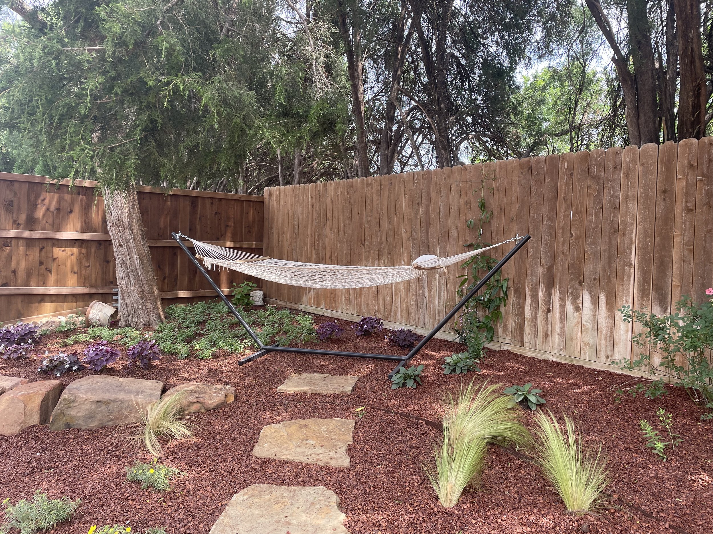 Backyard garden with a hammock between two trees, surrounded by plants, rocks, and a wooden fence.
