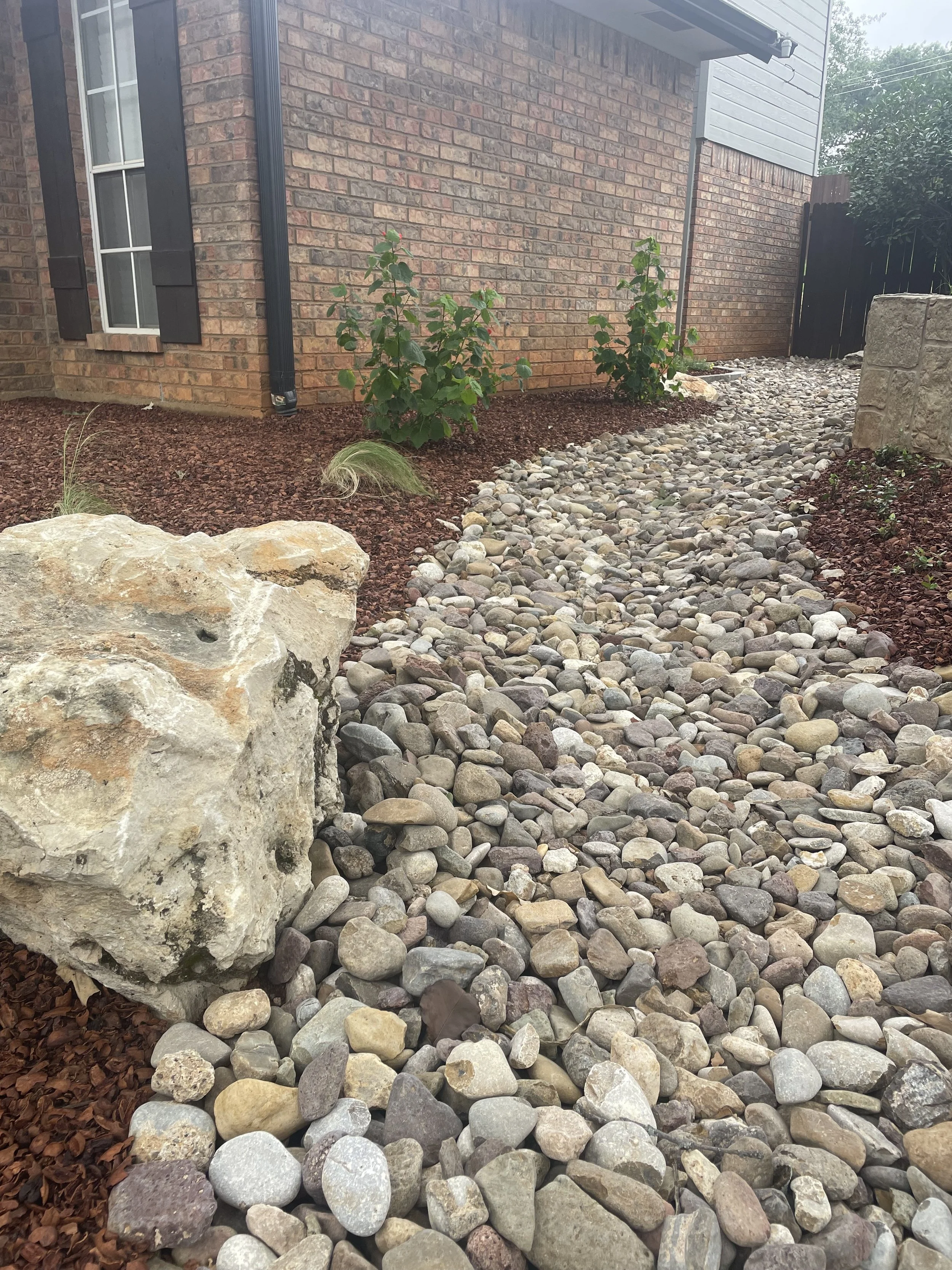 A gravel walkway with small stones, bordered by red mulch and two small bush plants, next to a brick house with a window and black shutters.