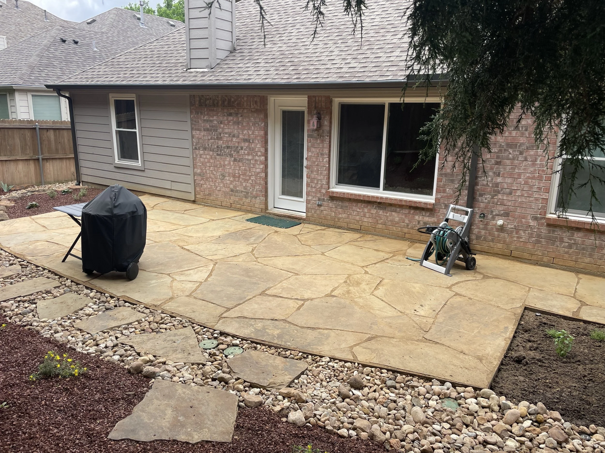 Newly installed stone patio in backyard with a barbecue grill covered with a black cover, a garden hose reel, a small garden bed with plants, and a pathway with stepping stones leading to the patio. The house has brick and siding exterior walls, and 