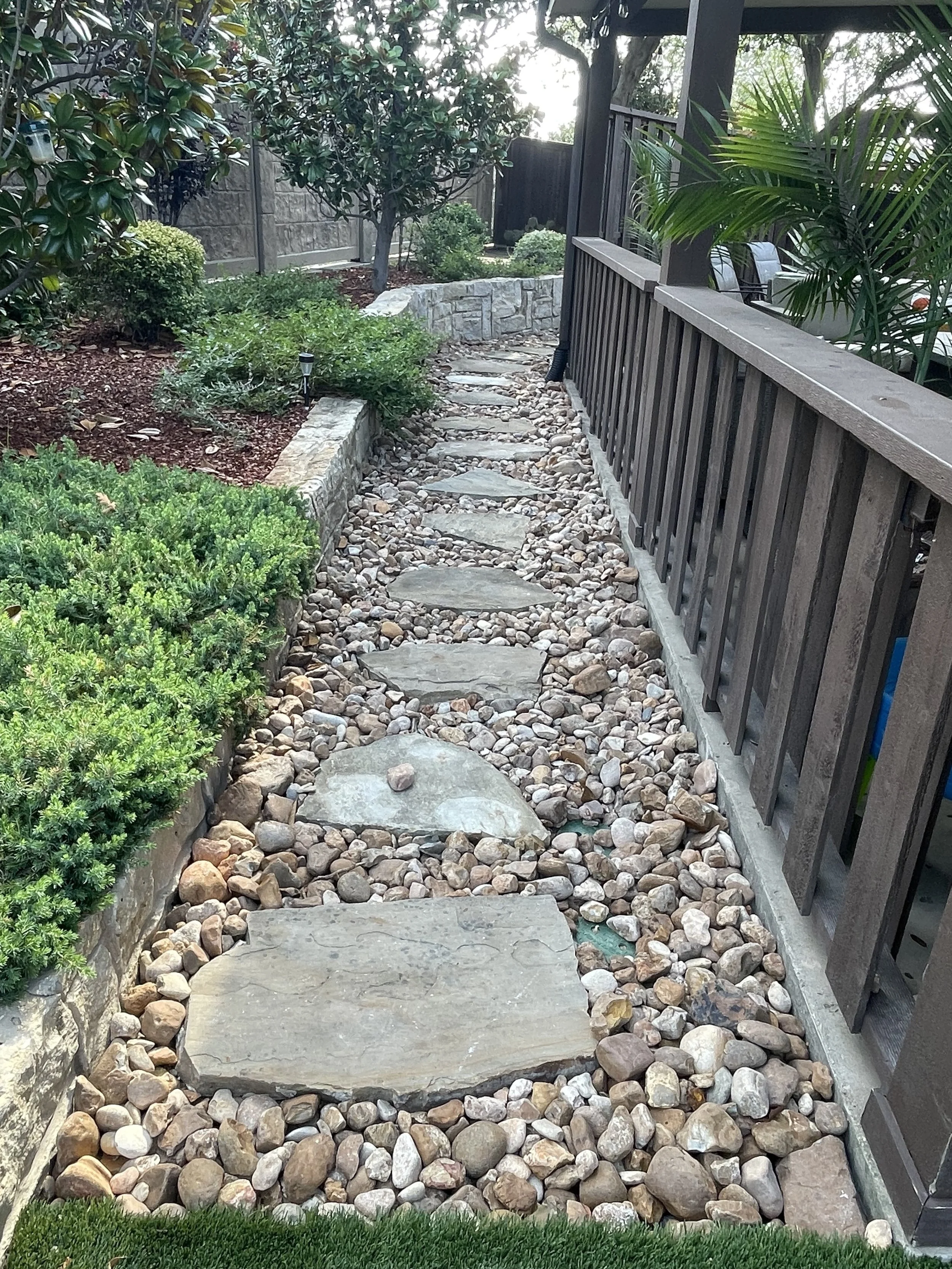 A narrow outdoor pathway made of large flat stones embedded in small rounded pebbles, bordered by a landscaped garden with various green bushes and trees, next to a wooden fence and porch.