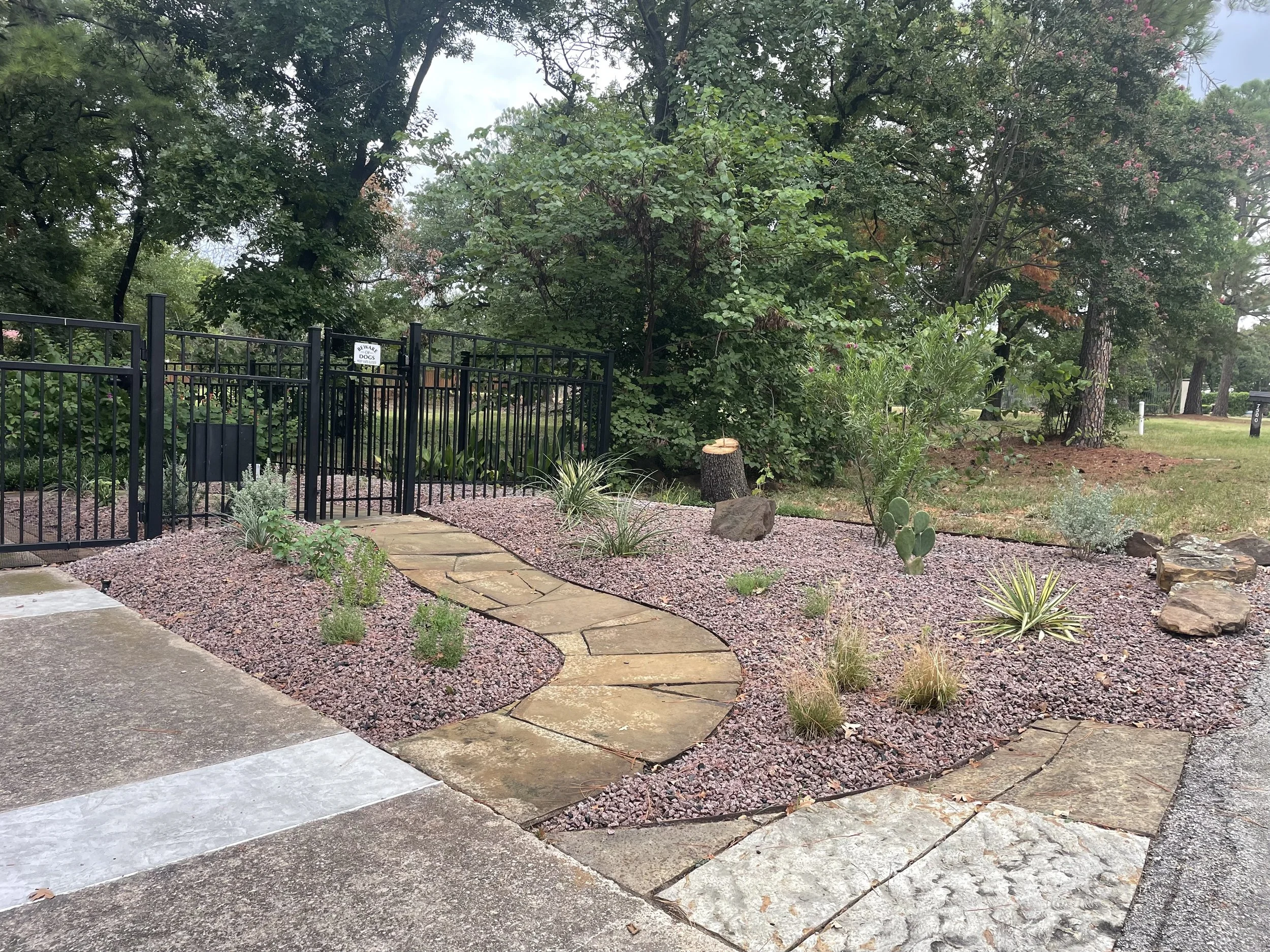 A garden with a winding stone pathway, surrounded by decorative rocks and desert plants, and enclosed by a black metal fence.