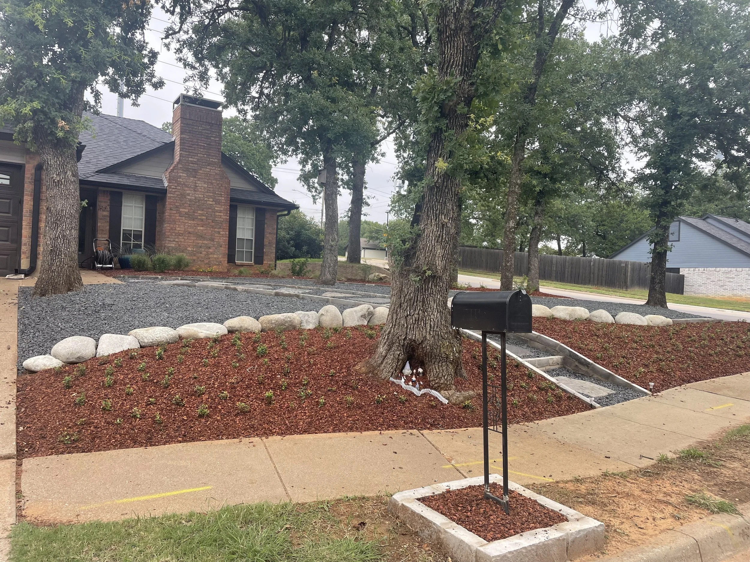 A residential front yard under landscaping with mulch, rocks, and small plants, with a brick house in the background and a black mailbox in the foreground.