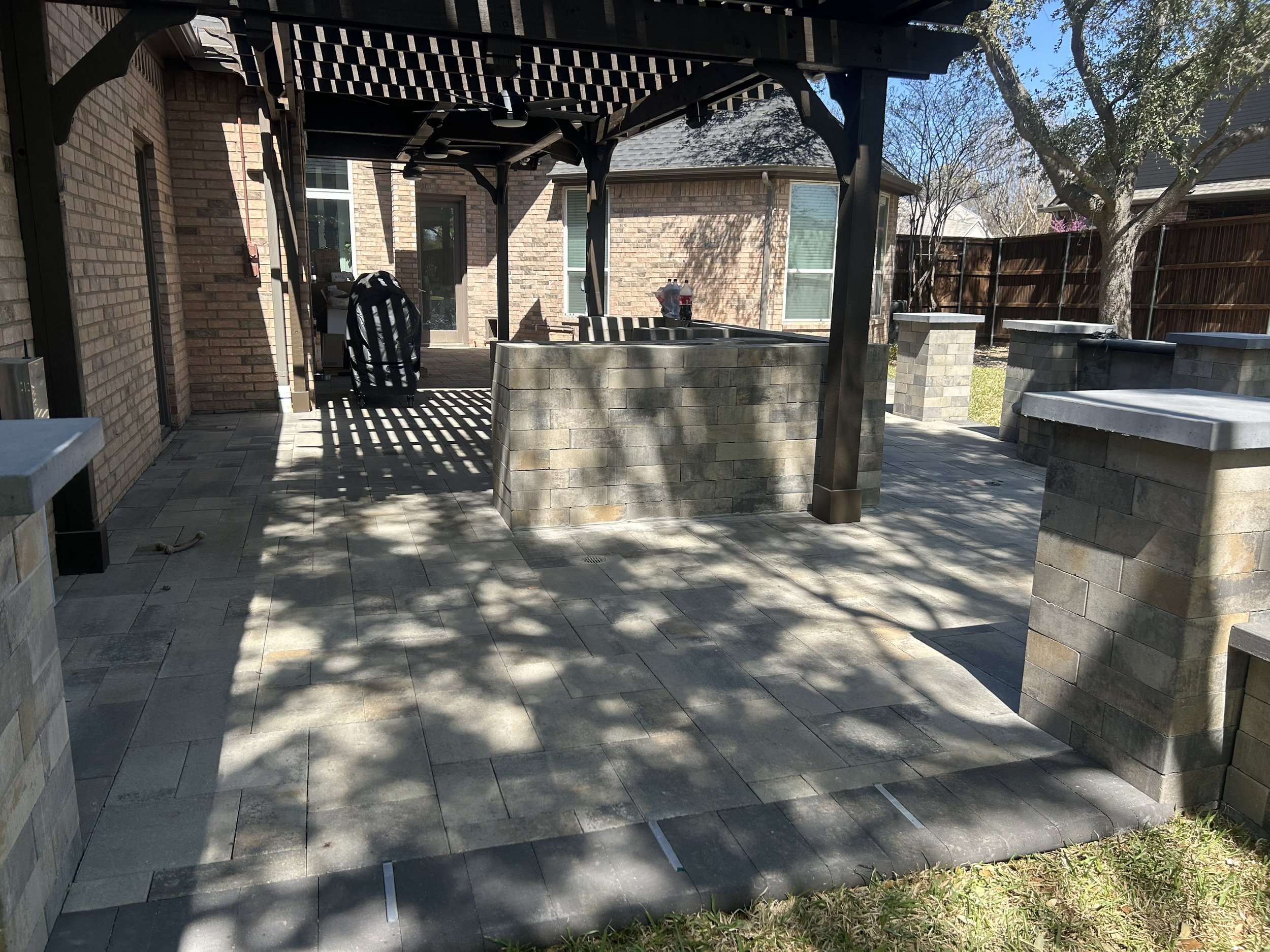 Backyard patio with stone flooring and brick house, shaded areas with trees, outdoor furniture, and a wooden fence.