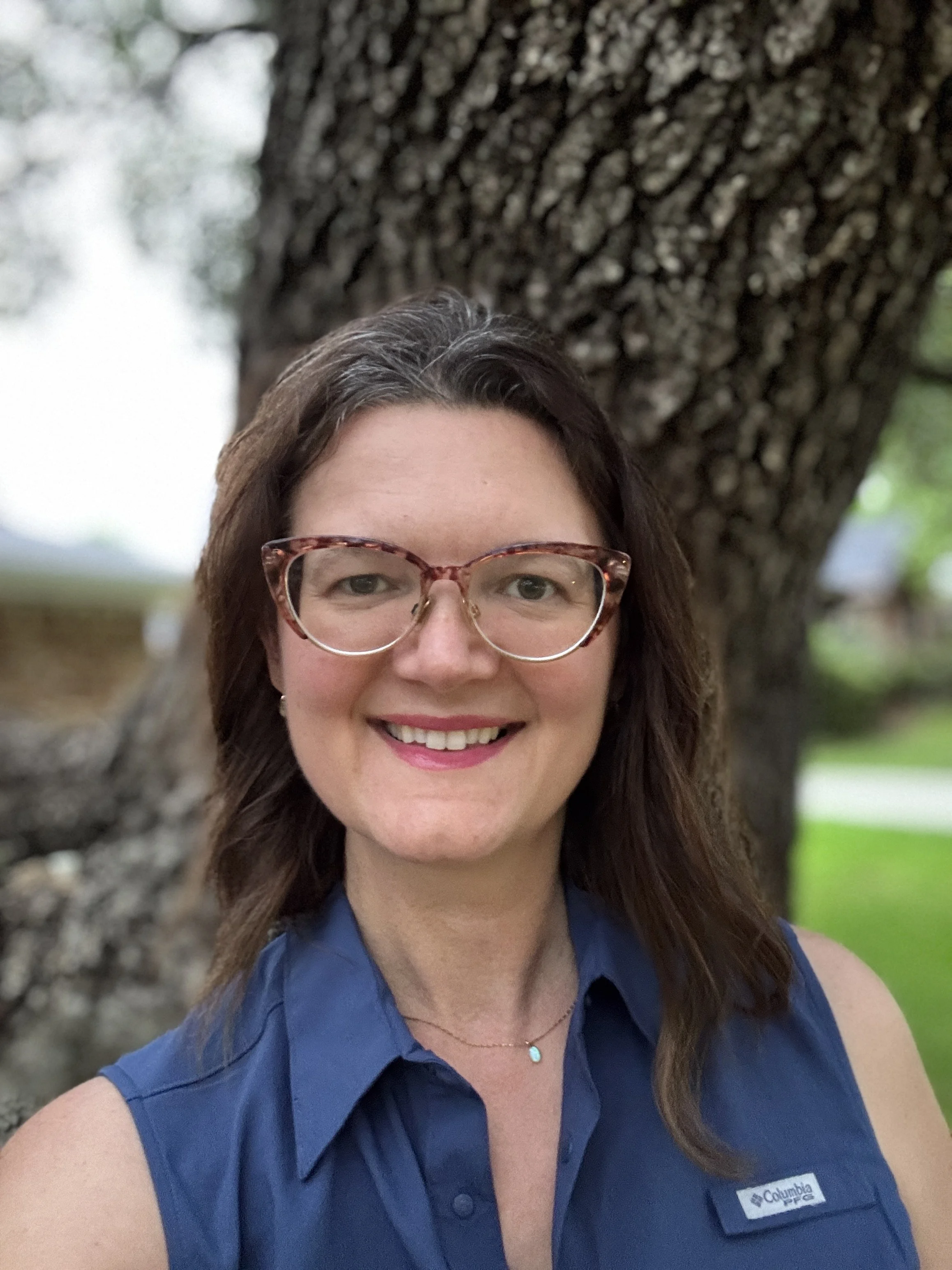 A woman with shoulder-length brown hair, wearing large tortoiseshell glasses, smiling, standing outdoors in front of a large tree trunk, wearing a blue Columbia vest and a delicate necklace with a small blue pendant.