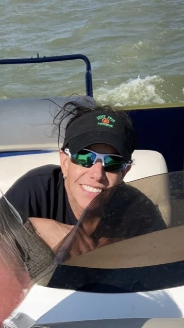 Woman wearing sunglasses, a black cap, and a black shirt on a boat, smiling at the camera, with water in the background.