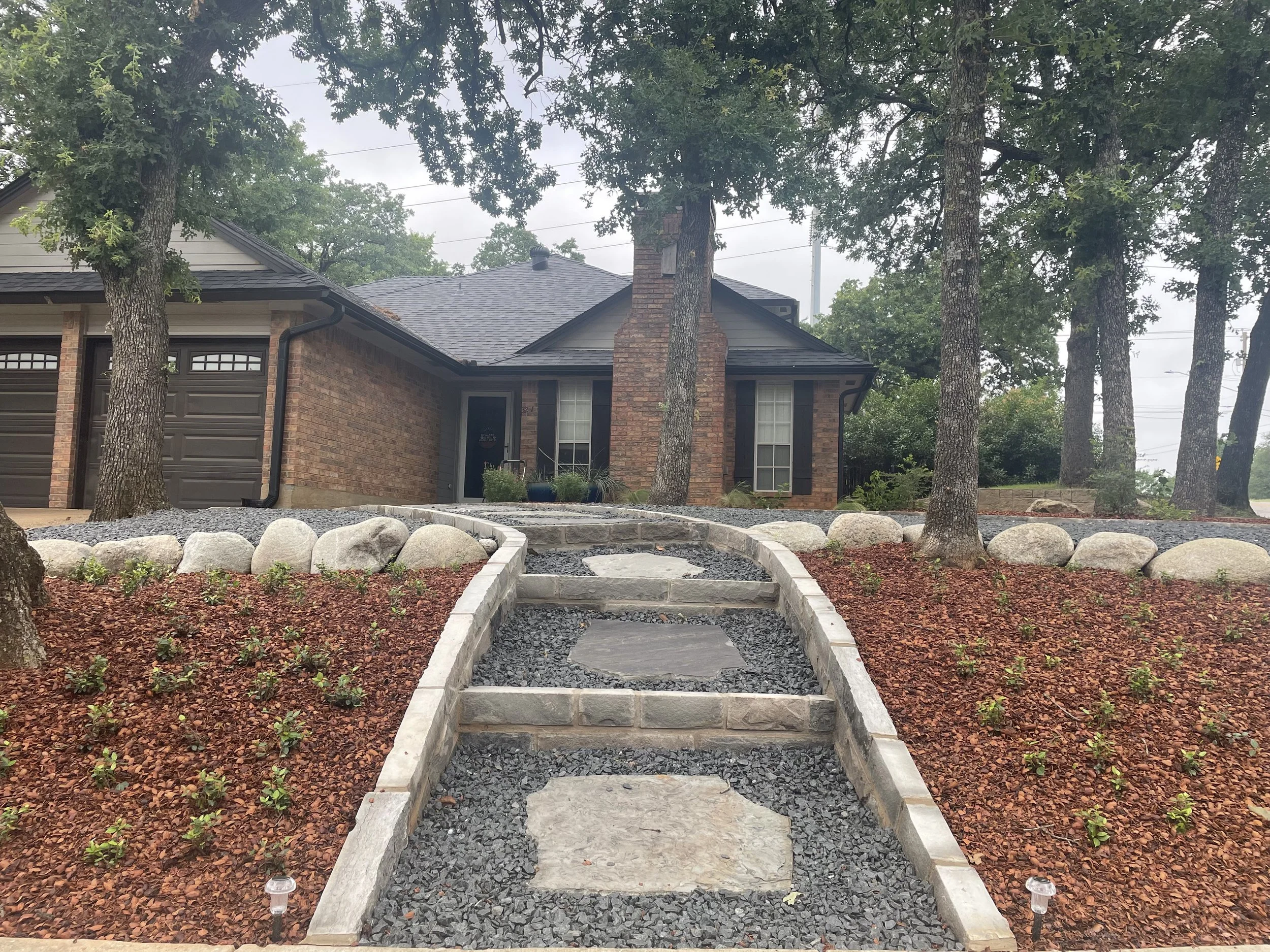 Front view of a brick house with a black roof, surrounded by trees and landscaped yard with a stone pathway and rocks.