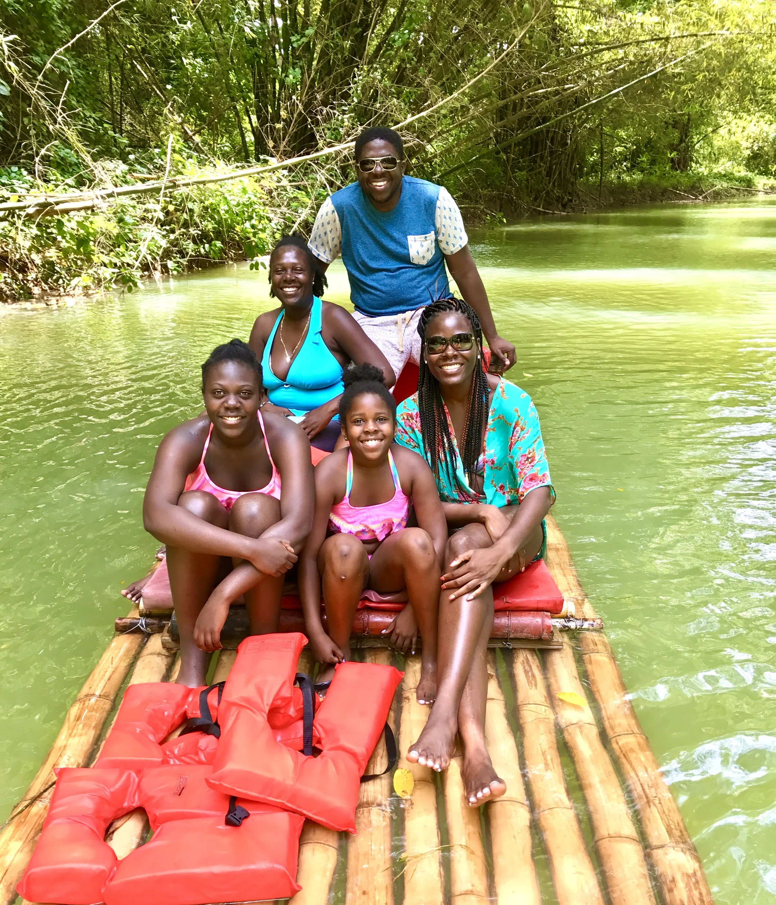 Group of people smiling and sitting on a bamboo raft on a river surrounded by green trees.