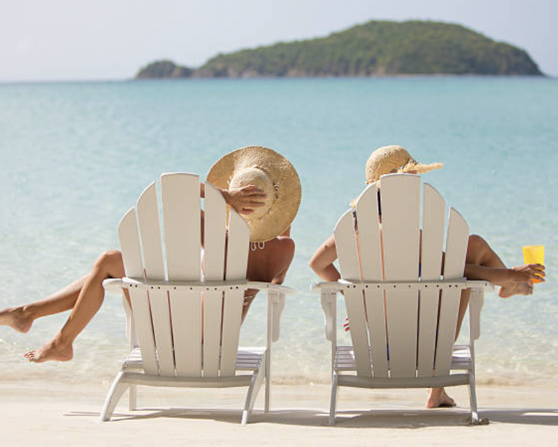 Two women wearing large sun hats sitting on white Adirondack chairs on the beach, with one in a bikini, at a tropical destination with a small island in the background, holding drinks.