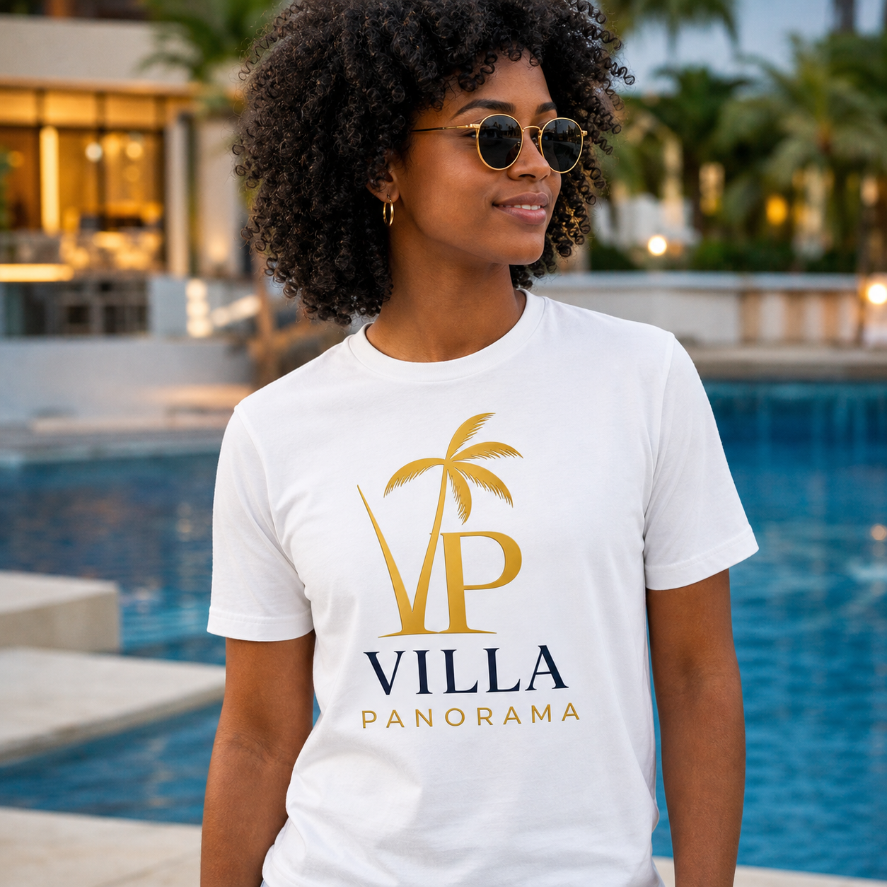 A woman wearing sunglasses and a white T-shirt with gold and black text and a palm tree graphic stands outdoors near a pool, with trees and a building in the background.