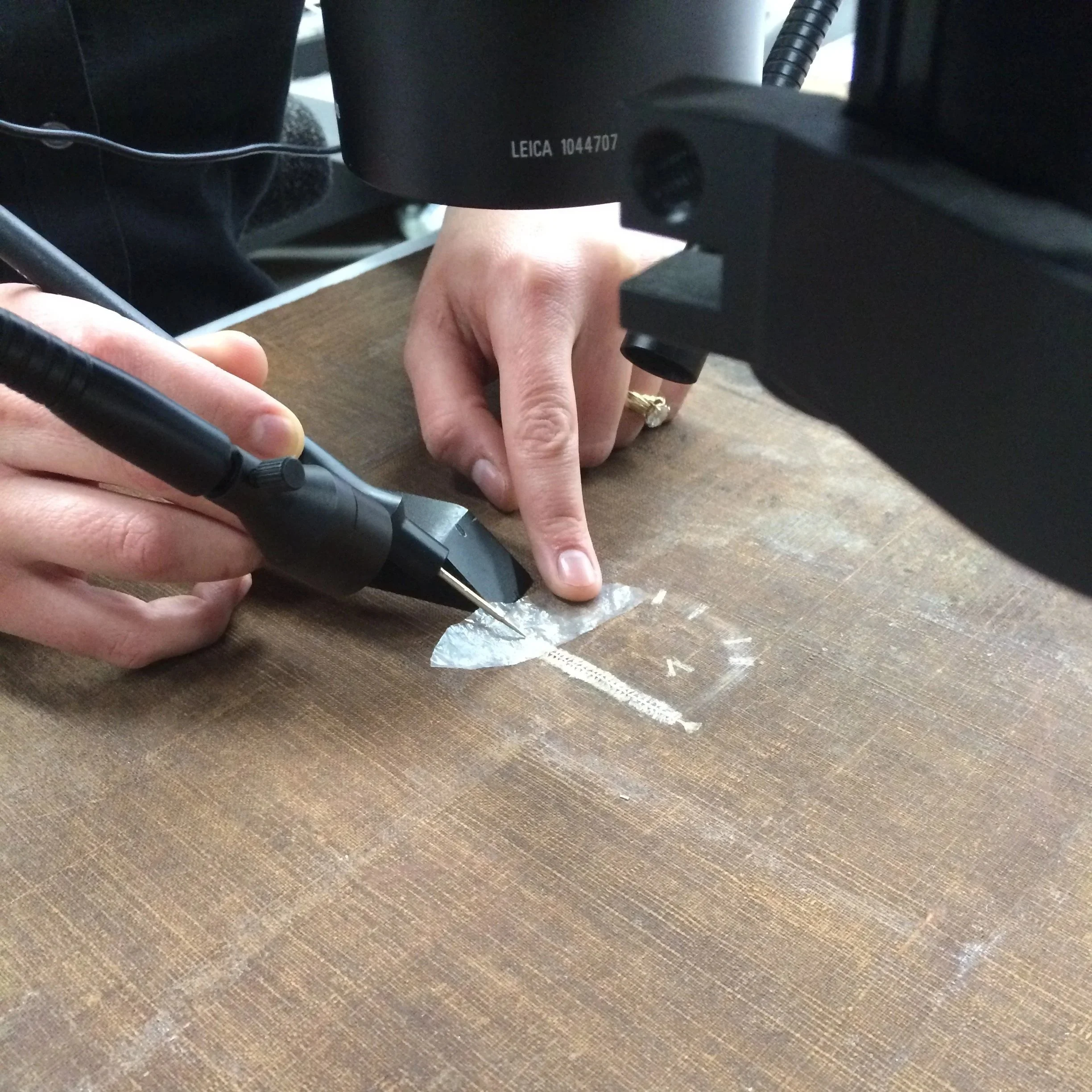 Close-up of a camera with the brand 'Leica' and serial number '1044707' visible, placed on a wooden surface, with a person's hand and a ring visible near the camera.