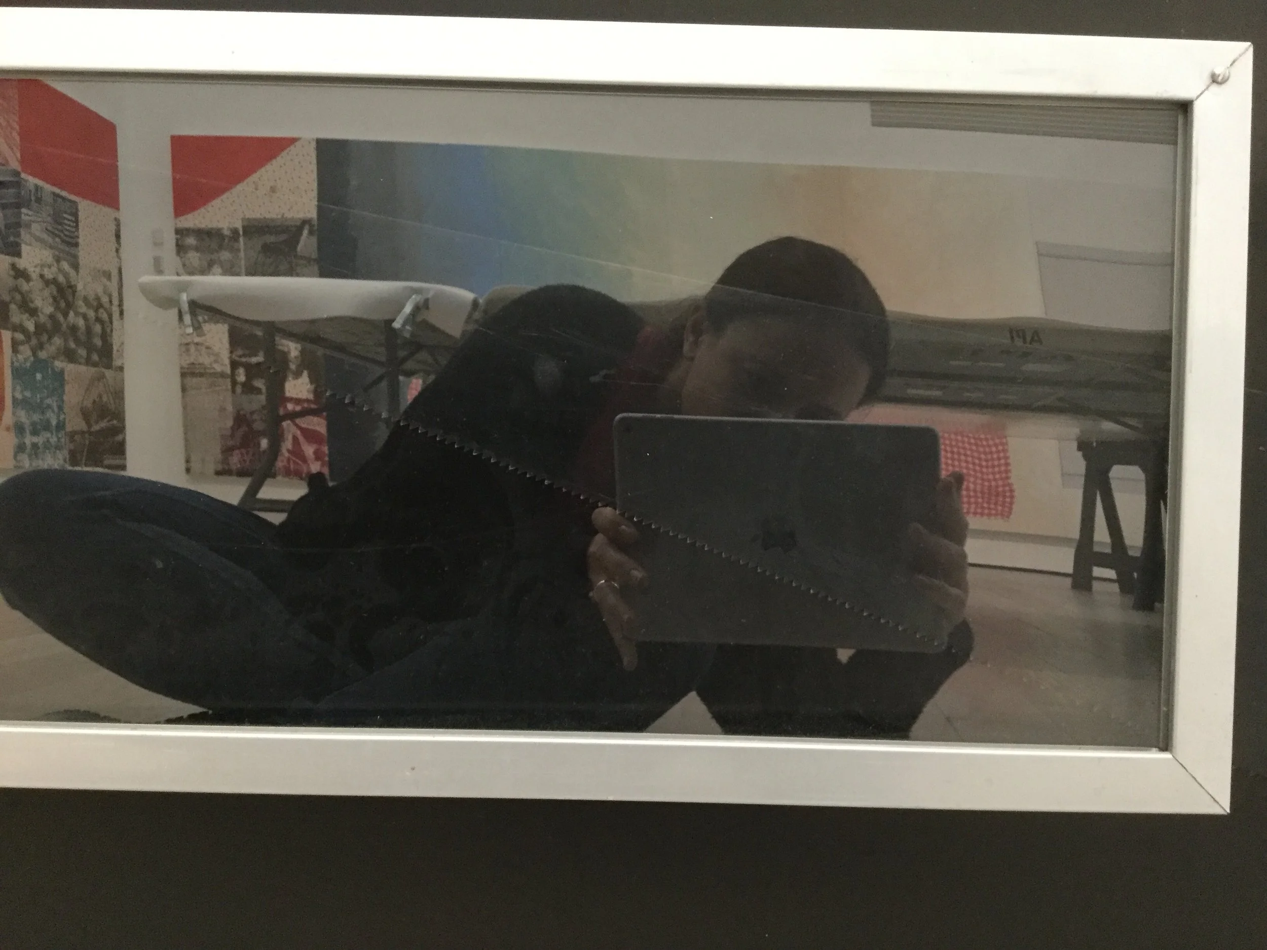 A person taking a photo of a mirror reflecting a room with a table and wall art.