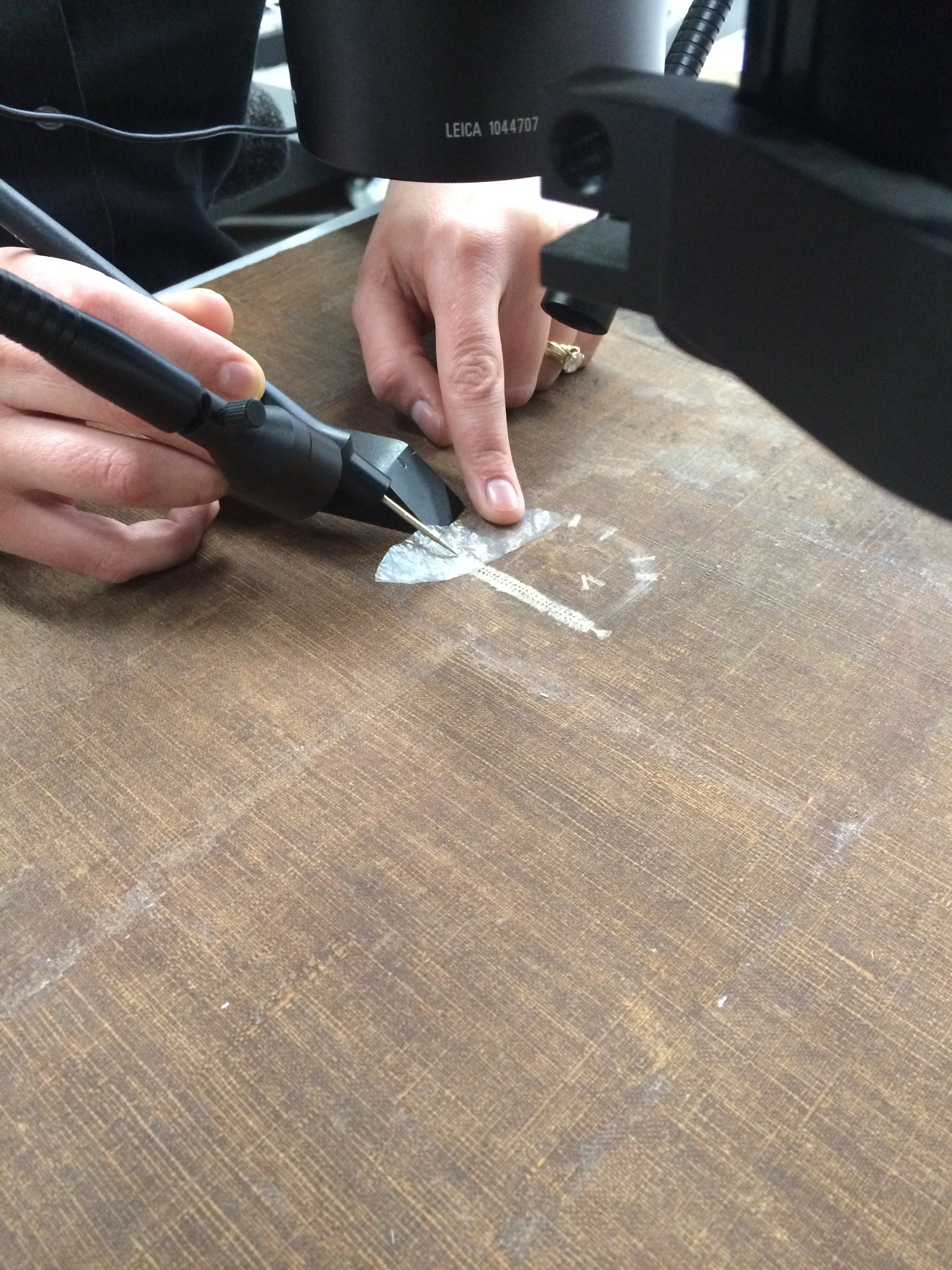 Close-up of a person's hand operating a soldering iron on a wooden surface, with a microscope above.