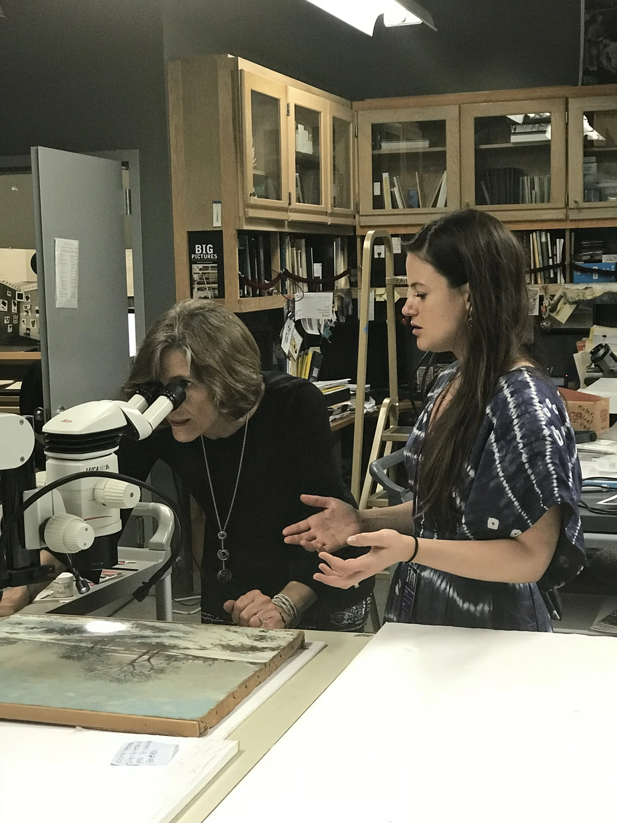 Two women are examining a specimen or object using a microscope in a laboratory or research setting, with shelves filled with books and files in the background.